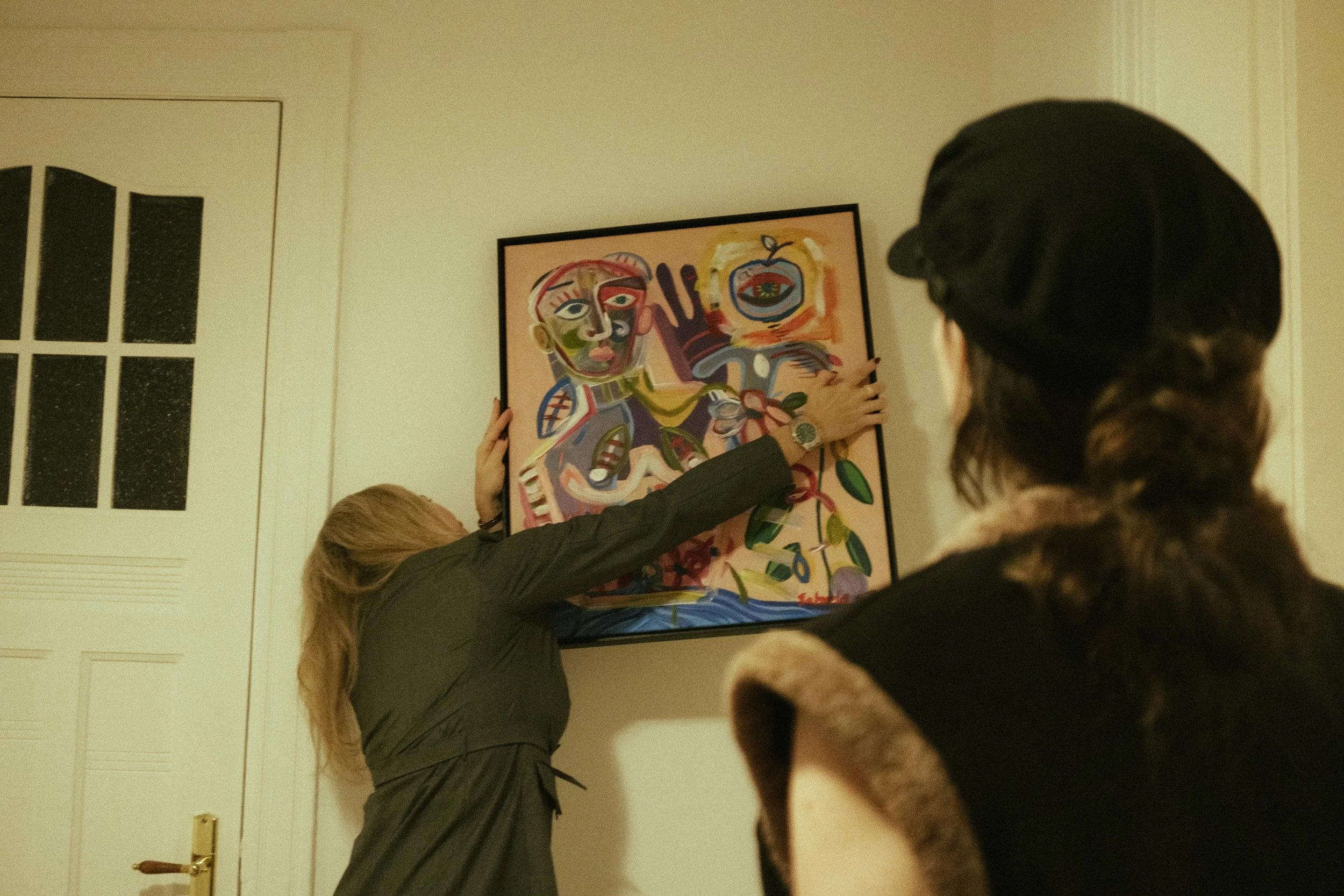 Two women observe a woman hanging a colorful abstract painting on a white wall.