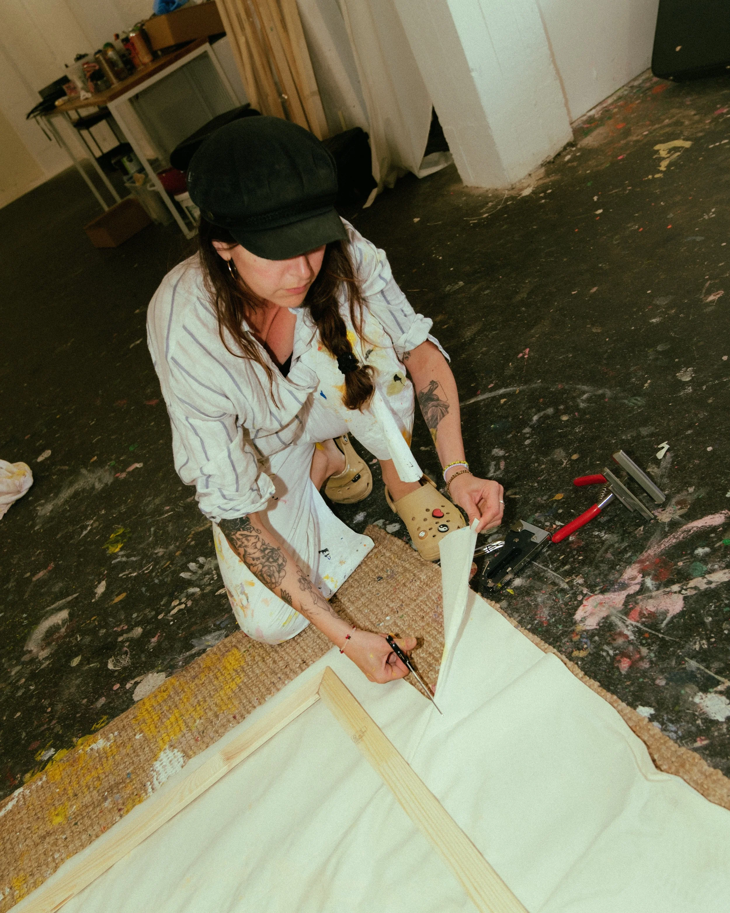 Woman with glasses, tattoos, and earrings working on a wood project in a room with a black floor, art supplies, and a white wall.