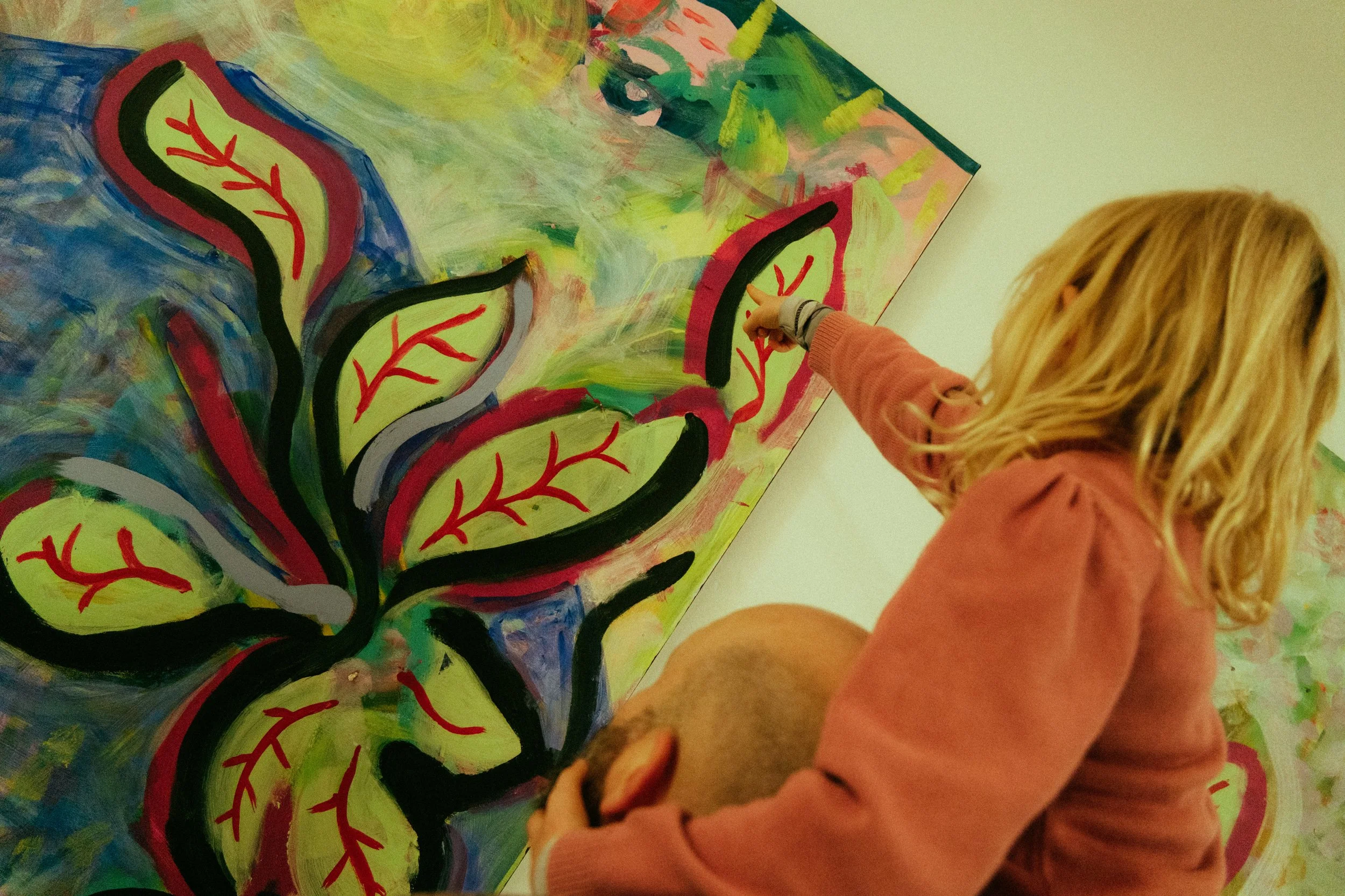 A young girl with blonde hair, wearing a pink sweatshirt, is sitting and pointing at a colorful abstract painting of a plant with red and green leaves on a canvas.