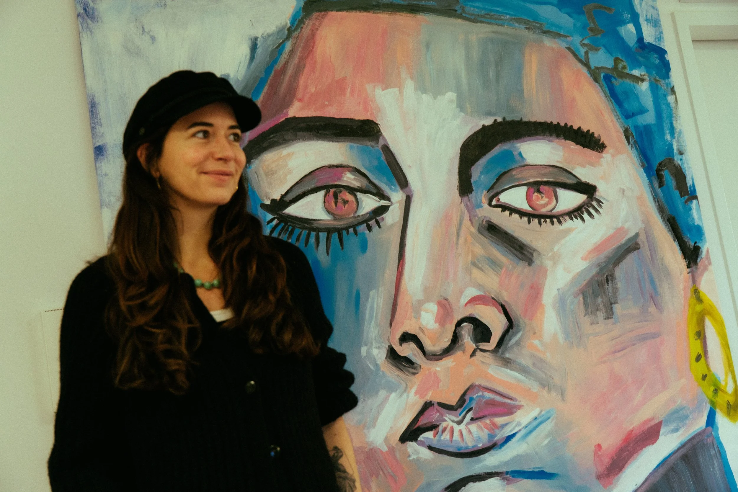 A woman with long brown hair wearing a black hat and black sweater standing next to a large colorful abstract painting of a woman's face with pink eyes, bold eyebrows, and yellow earrings.