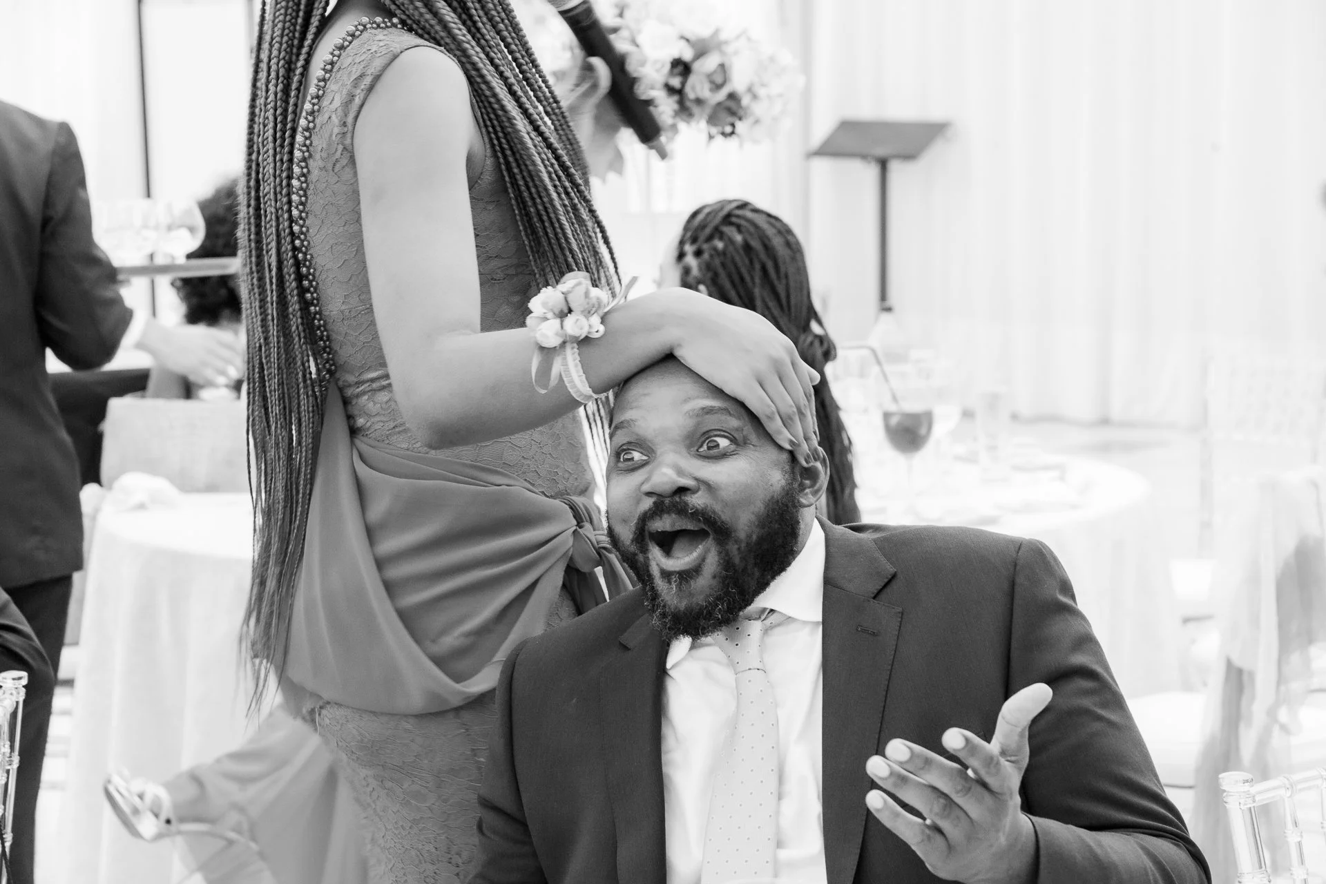 Black and white photo of a woman with long braids and a wrist corsage playfully touching a man's head, who is smiling and gesturing with his hand. They are at a reception with tables, drinks, and people in the background.