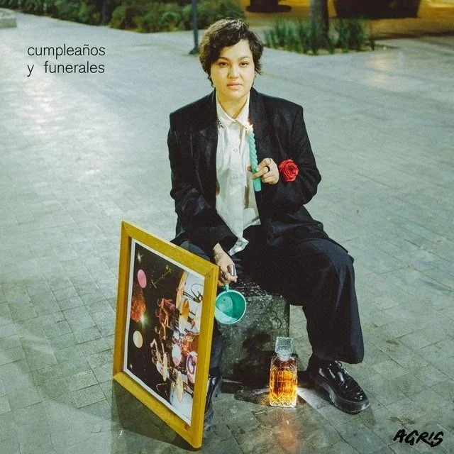 A woman sitting on a concrete block on a street at night, holding a cigarette and a red rose. She is dressed in a black blazer and pants, with a white shirt. There are birthday and funeral items around her, including a framed picture of a space scene, a small blue bucket, and a bottle of whiskey. The words "cumpleaños y funerales" are written on the image, meaning "birthdays and funerals" in Spanish.