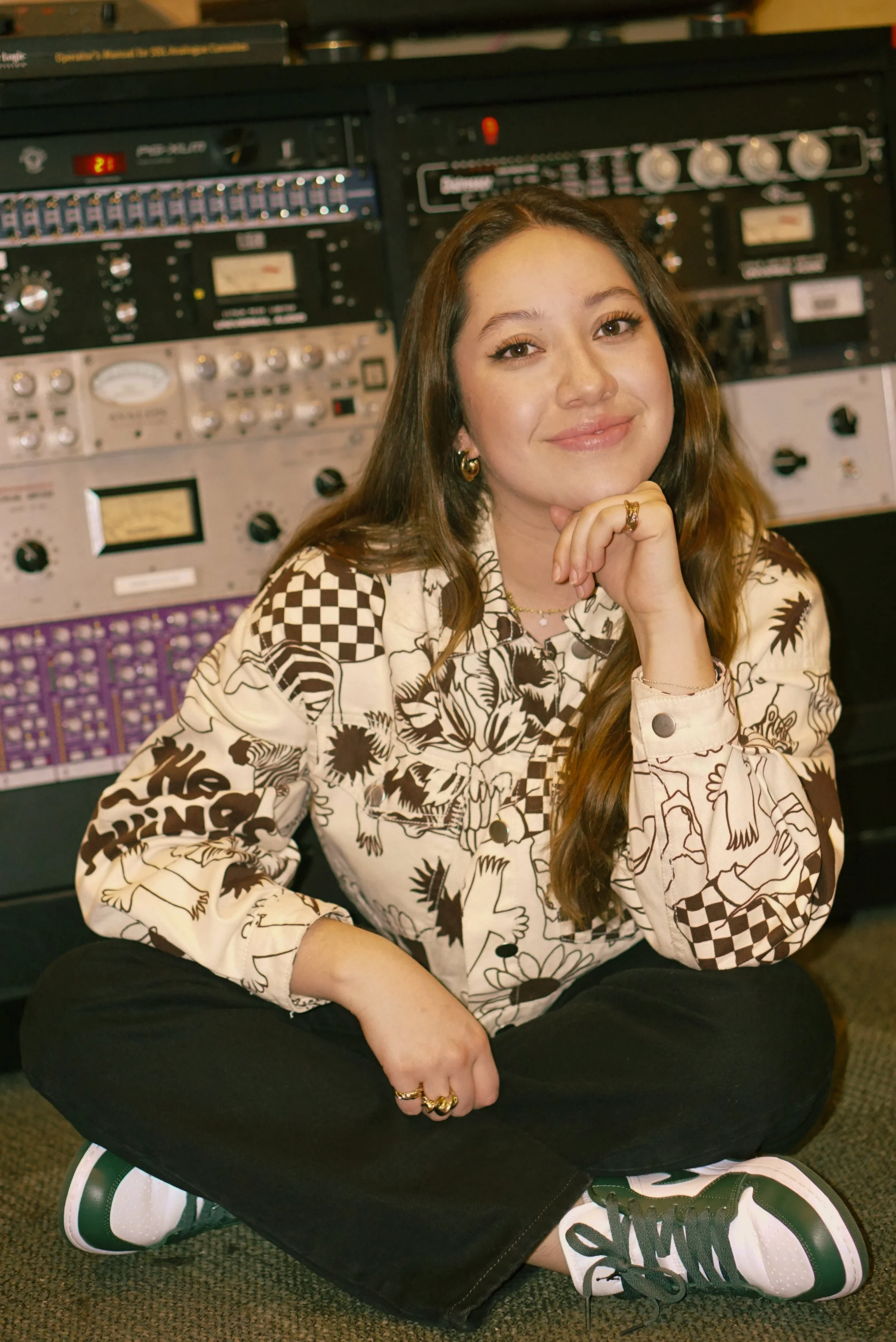 A young woman with long brown hair sitting cross-legged on the floor, smiling at the camera, in front of audio equipment.