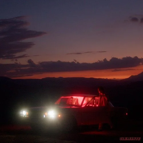 A person standing outside a car with red lights inside, parked on a dark landscape during sunset or dusk, with a dramatic sky and clouds in the background.