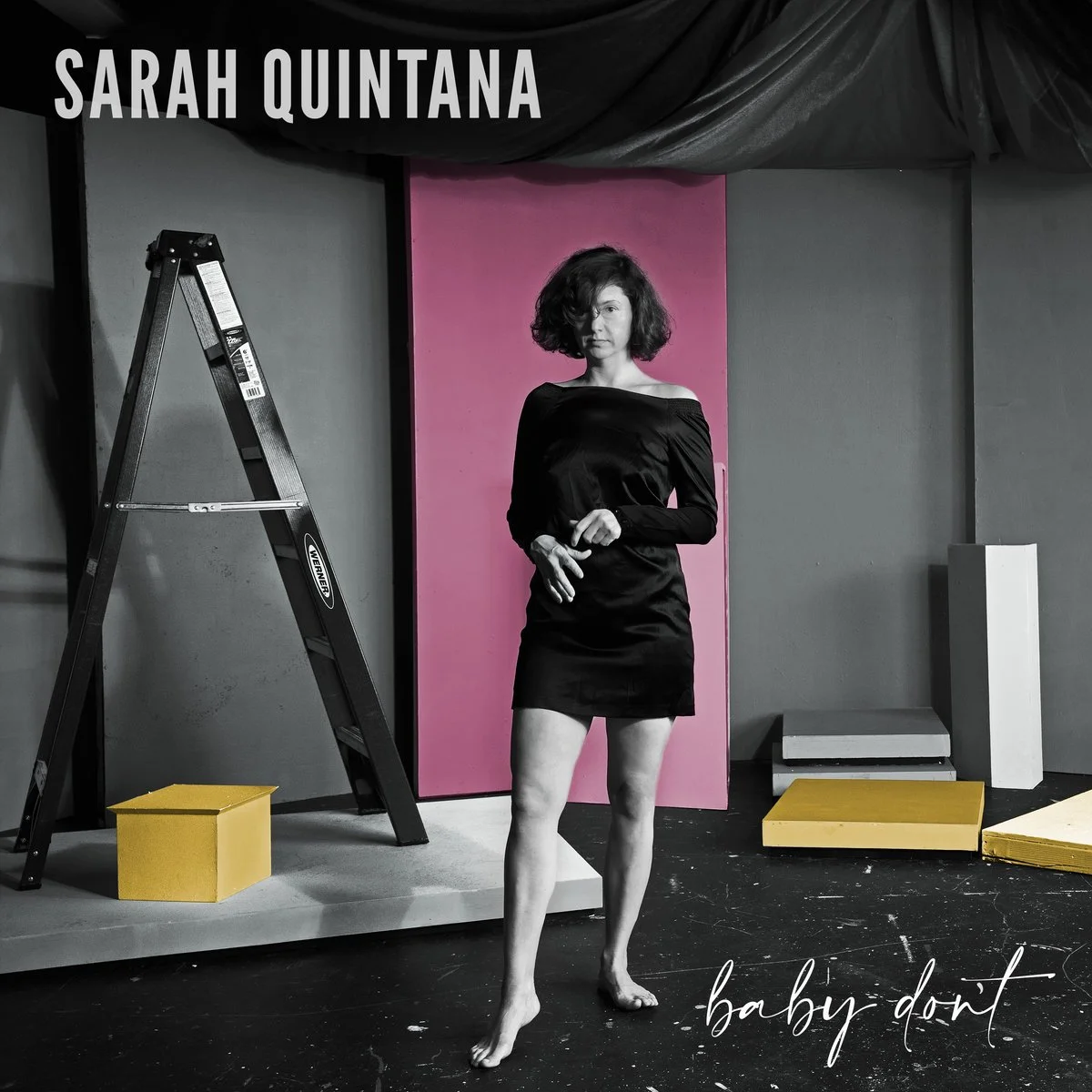 A woman with short curly hair standing barefoot in a studio with colorful geometric backgrounds. She wears a black off-the-shoulder dress, with one hand clutching her other wrist. The studio contains a ladder, a yellow box, and several colorful blocks. The text 'Sarah Quintana' is in the top left corner, and 'baby don't' is in cursive at the bottom right.