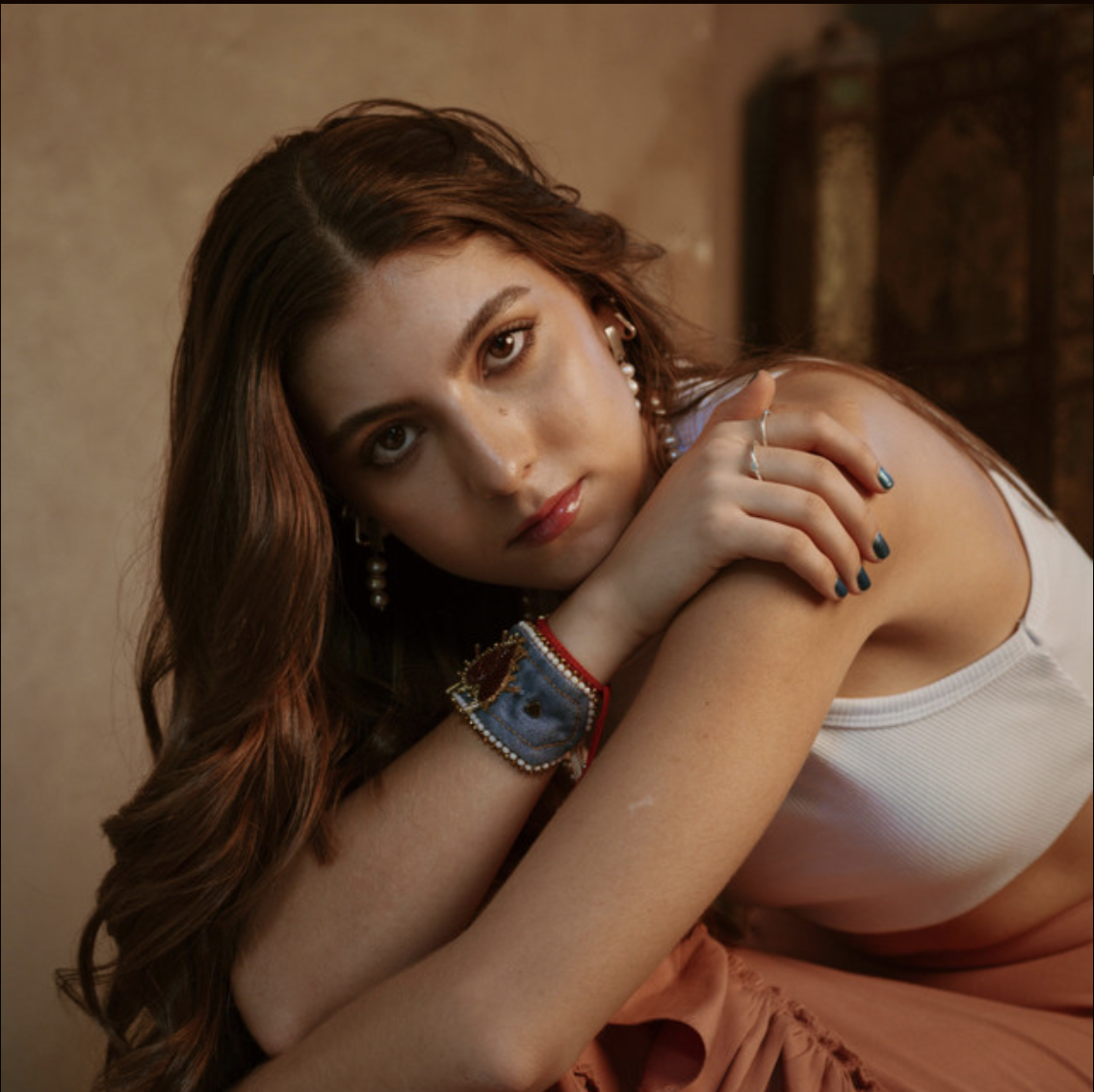 A young woman with long wavy brown hair, wearing a white sleeveless top and peach-colored skirt, rests her chin on her hand. She is decorated with earrings, a bracelet, and rings, and has her nails painted dark blue. The background is blurred with warm tones.
