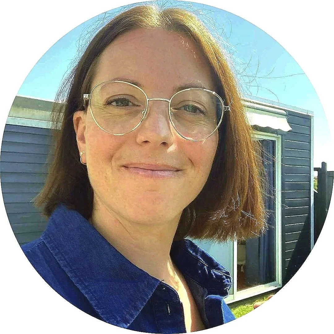 En kvinde med briller, skulderlangt brunt hår, blå skjorte, smilende, udendørs med blå bygning og himmel i baggrunden. Terapeuten Denise Thinggaard