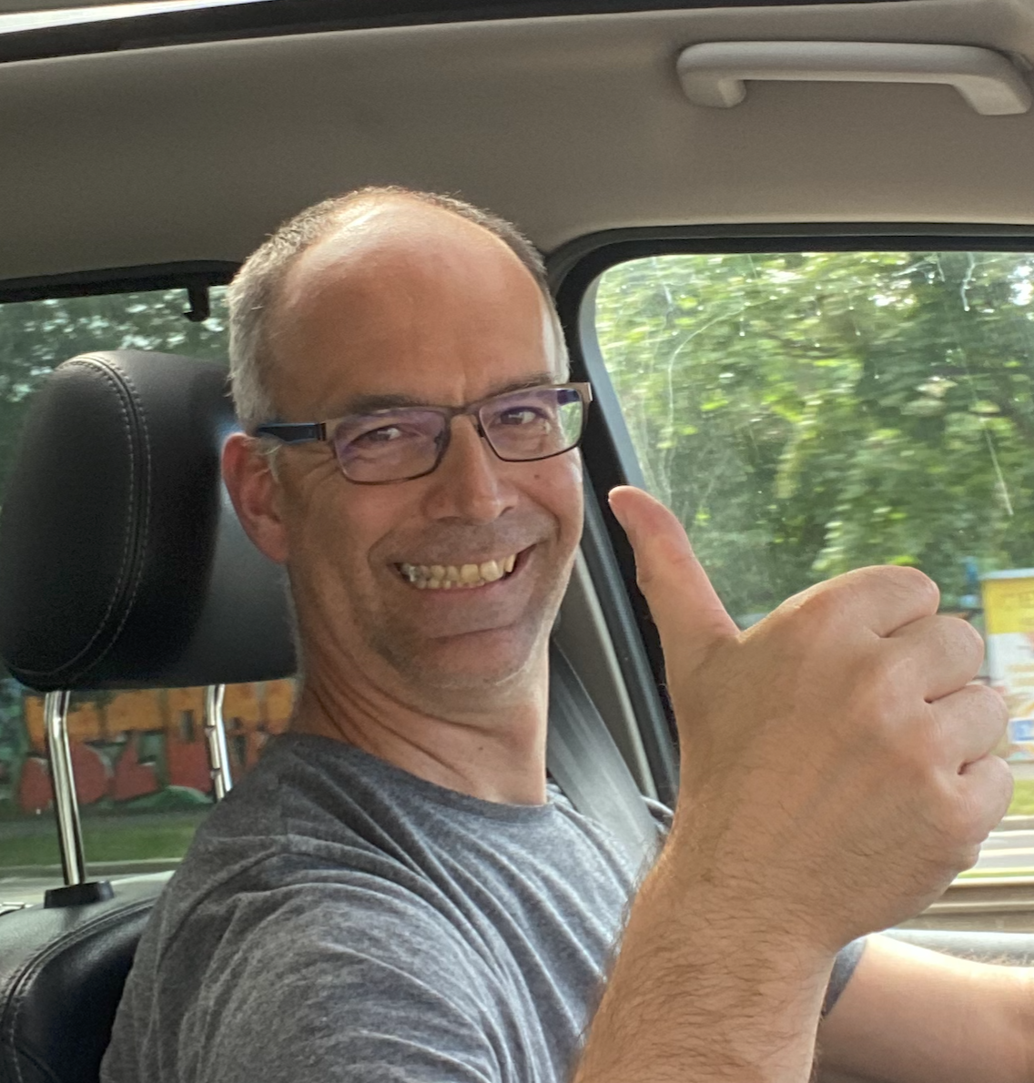 A smiling man wearing glasses giving a thumbs-up inside a vehicle, with green trees outside the window.