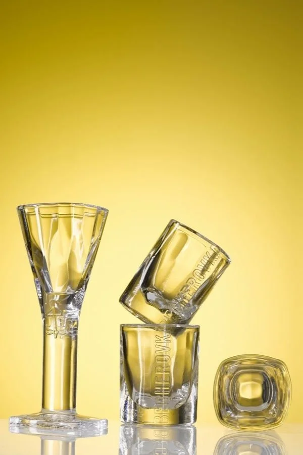 Four transparent shot glasses with the Tesoro logo, one filled with a yellow liquid, one tilted, one stacked, and one on its side, against a yellow background.