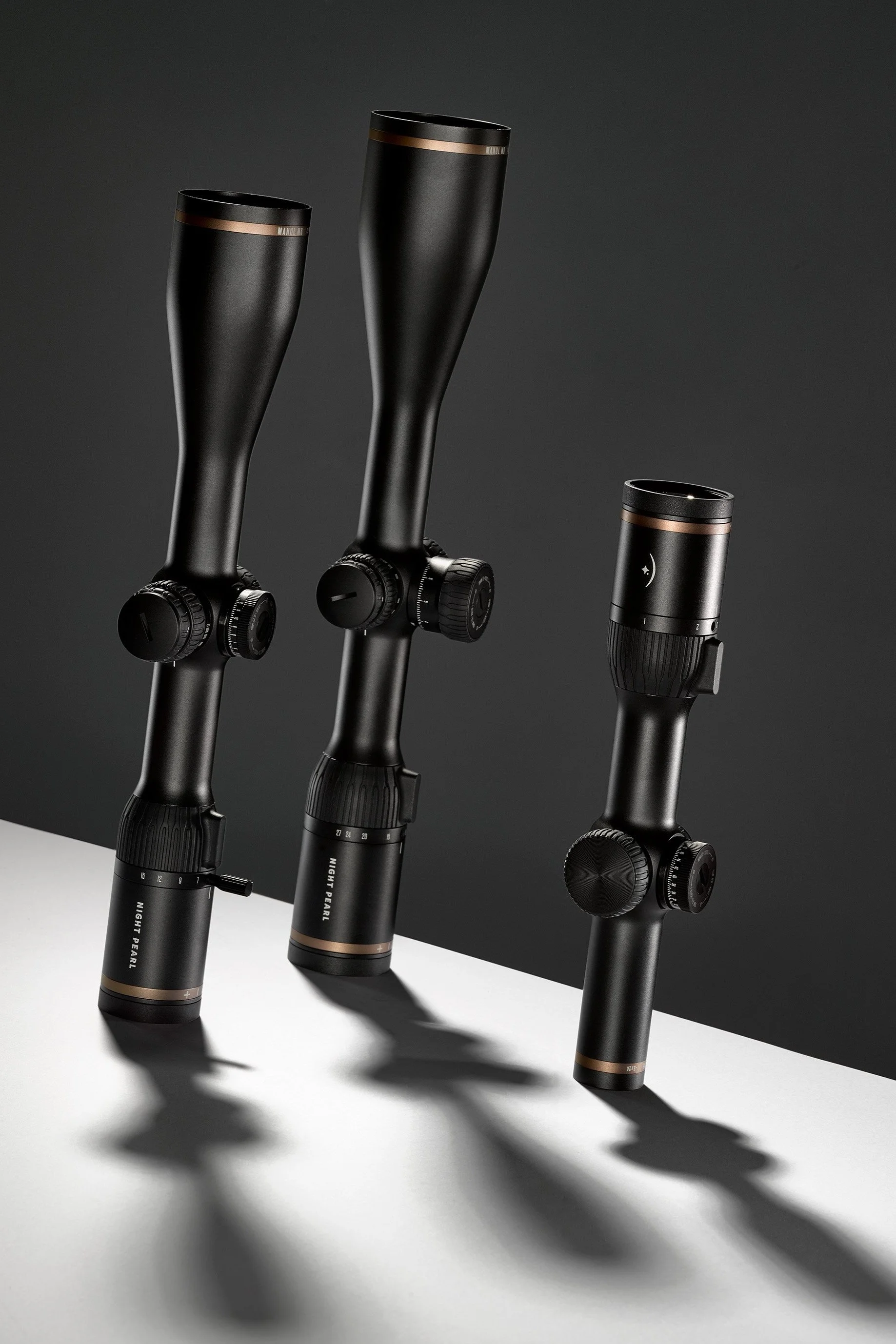 Three black rifle scopes with orange accents on a white surface with a dark background, casting shadows.