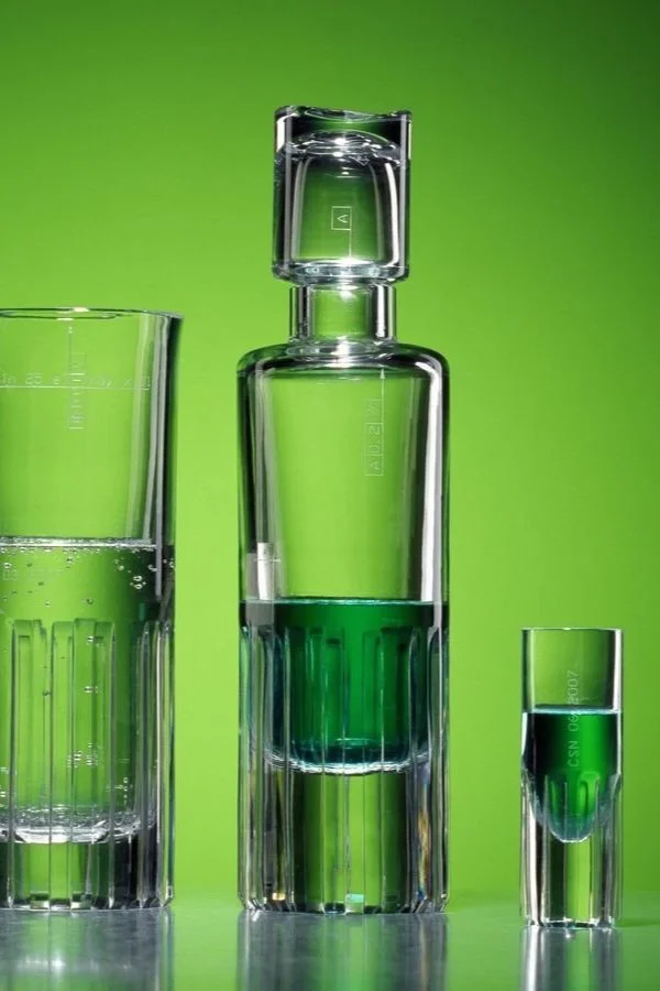 A set of three glass containers with green liquid against a bright green background. The largest container is a tall cylindrical bottle with a glass stopper, in the middle. To the left, a slightly shorter tall glass with measuring marks, and to the r