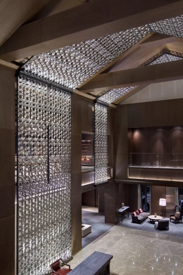 Elegant hotel lobby with decorative glass walls and wooden accents, featuring seating areas with chairs and tables.