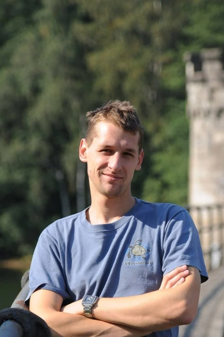 A young man with short brown hair, wearing a blue t-shirt with a cartoonish whale logo, standing outdoors with arms crossed, smiling at the camera.