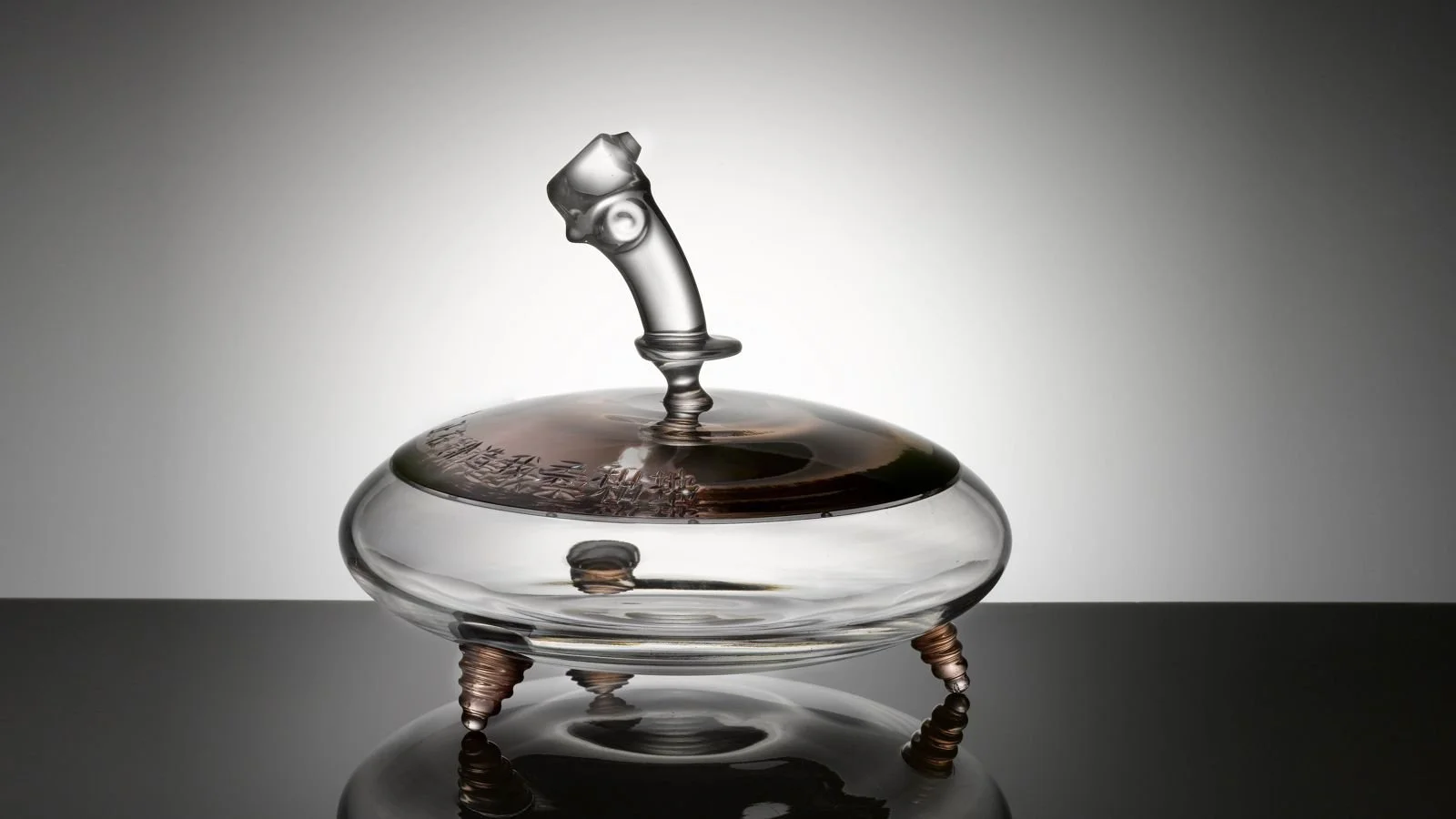 A glass perfume bottle with a uniquely shaped cap, set on a reflective black surface, with a neutral background.