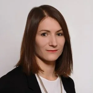 Portrait of a young woman with shoulder-length brown hair, wearing a black blazer over a white top, against a plain gray background.