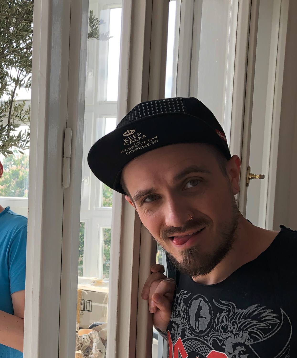 A man with a nose ring, beard, and mustache peeking through a door with a playful expression, wearing a black cap with text, and a black graphic t-shirt.