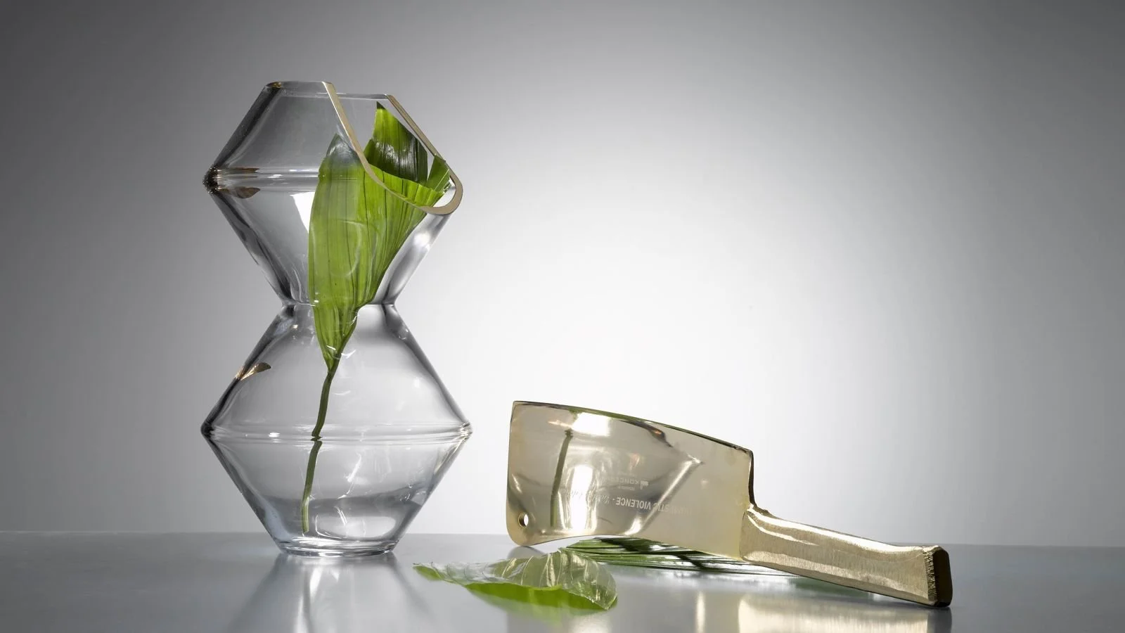 A broken glass vase with green leaves inside, and a large glass knife lying on its side, reflecting on a white surface against a gray background.