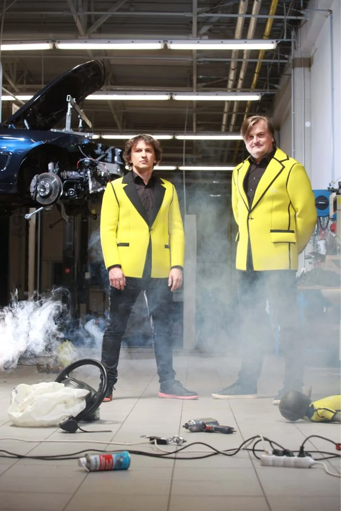 Two men wearing yellow jackets standing in a garage with a partially disassembled car in the background. There is smoke on the floor and various tools and objects around them, including a smoke canister and a vacuum.