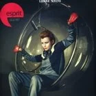 A person sitting in a spaceship-like capsule with a dark interior, wearing a blue outfit and a red glove on one hand, raising their arm.