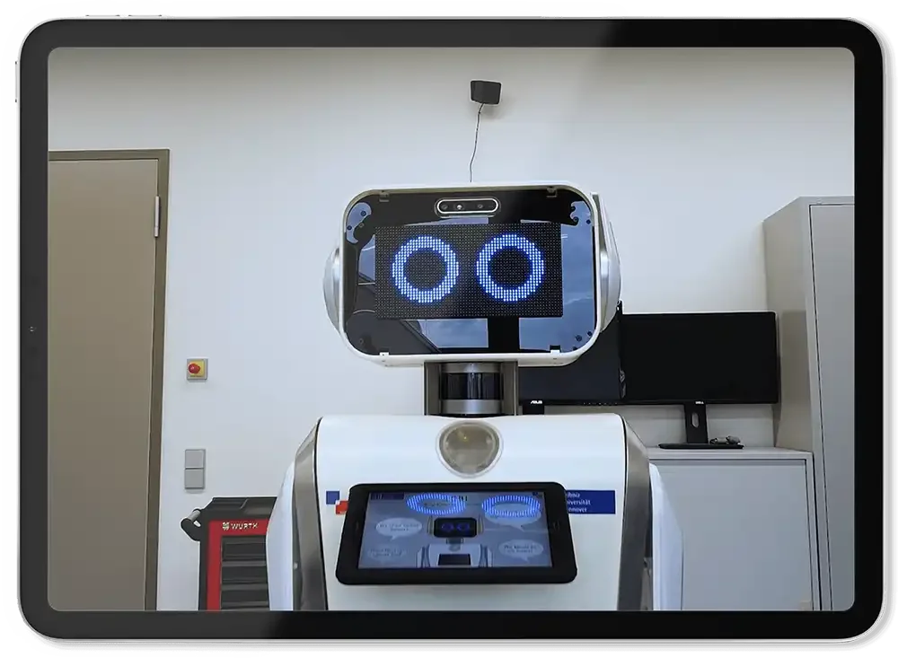 Ein humanoider Roboter mit einem Bildschirm im Kopf, der ein lachendes Gesicht zeigt, steht in einem Raum mit weißen Wänden, einem schwarzen Monitor, einem grauen Schrank und einem roten Werkzeugwagen.