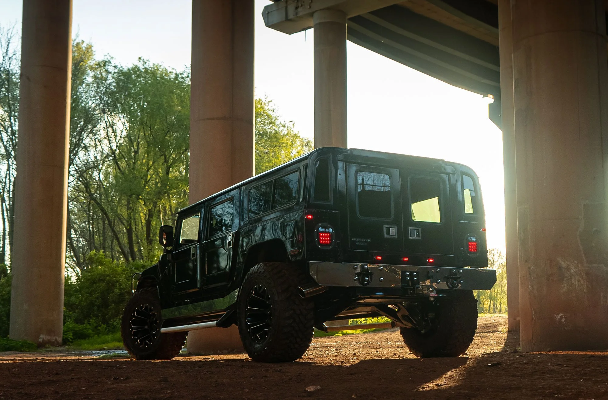 Black Hummer SUV parked under a bridge, surrounded by concrete pillars and trees in the background.