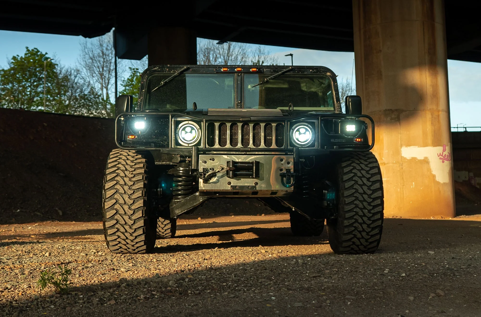 Front view of a modified military-style vehicle with large tires and bright headlights, parked under a bridge.