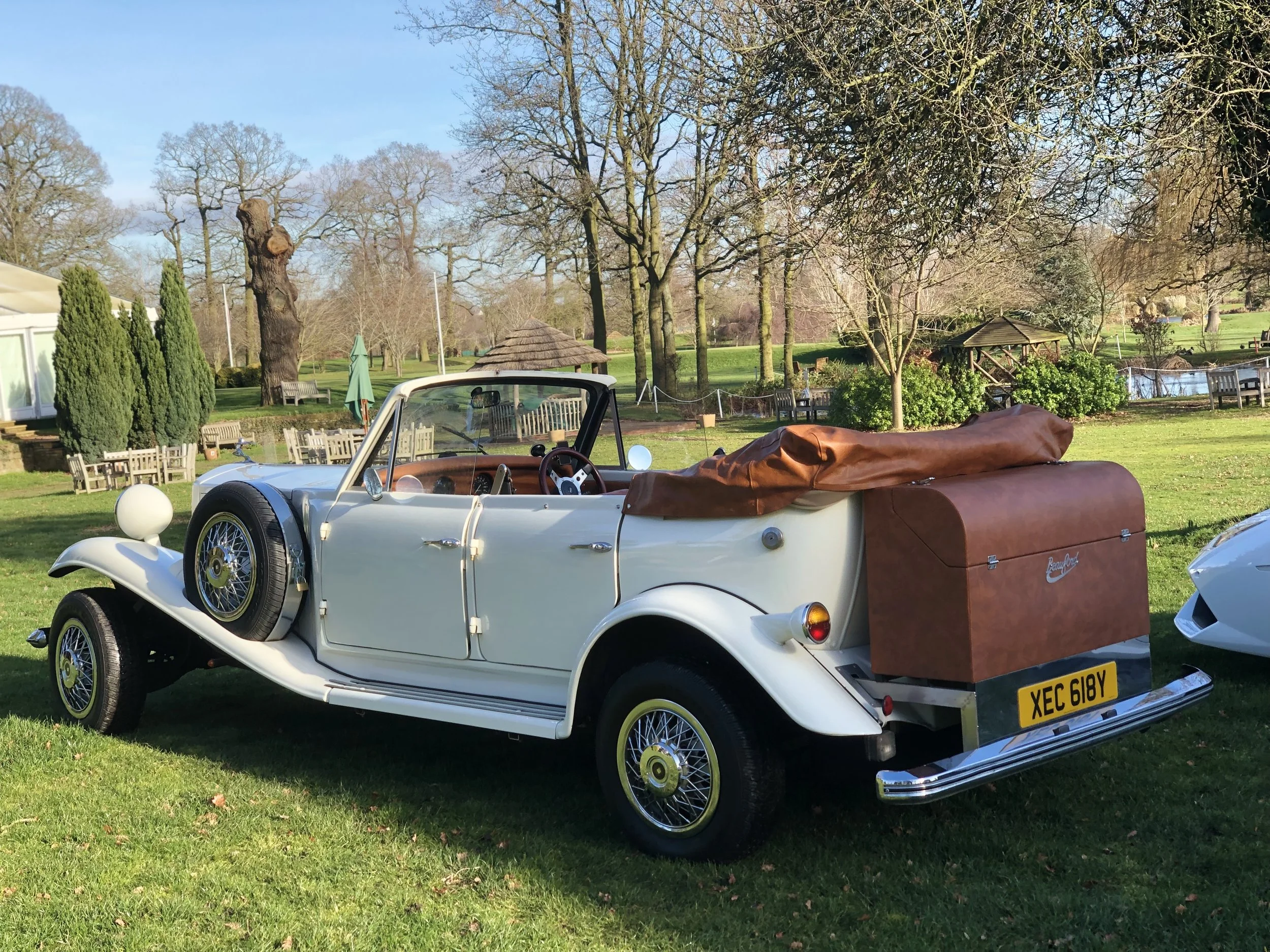 Vintage white convertible car with a brown leather-covered luggage trunk, parked on grass with trees and outdoor seating in the background.