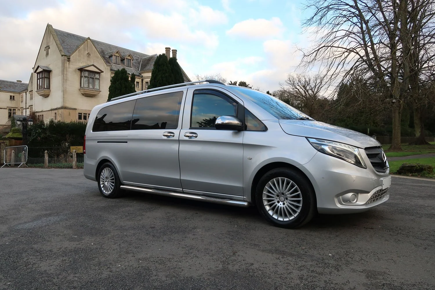 Silver Mercedes-Benz van parked in front of a large stone building with trees and a clear sky.