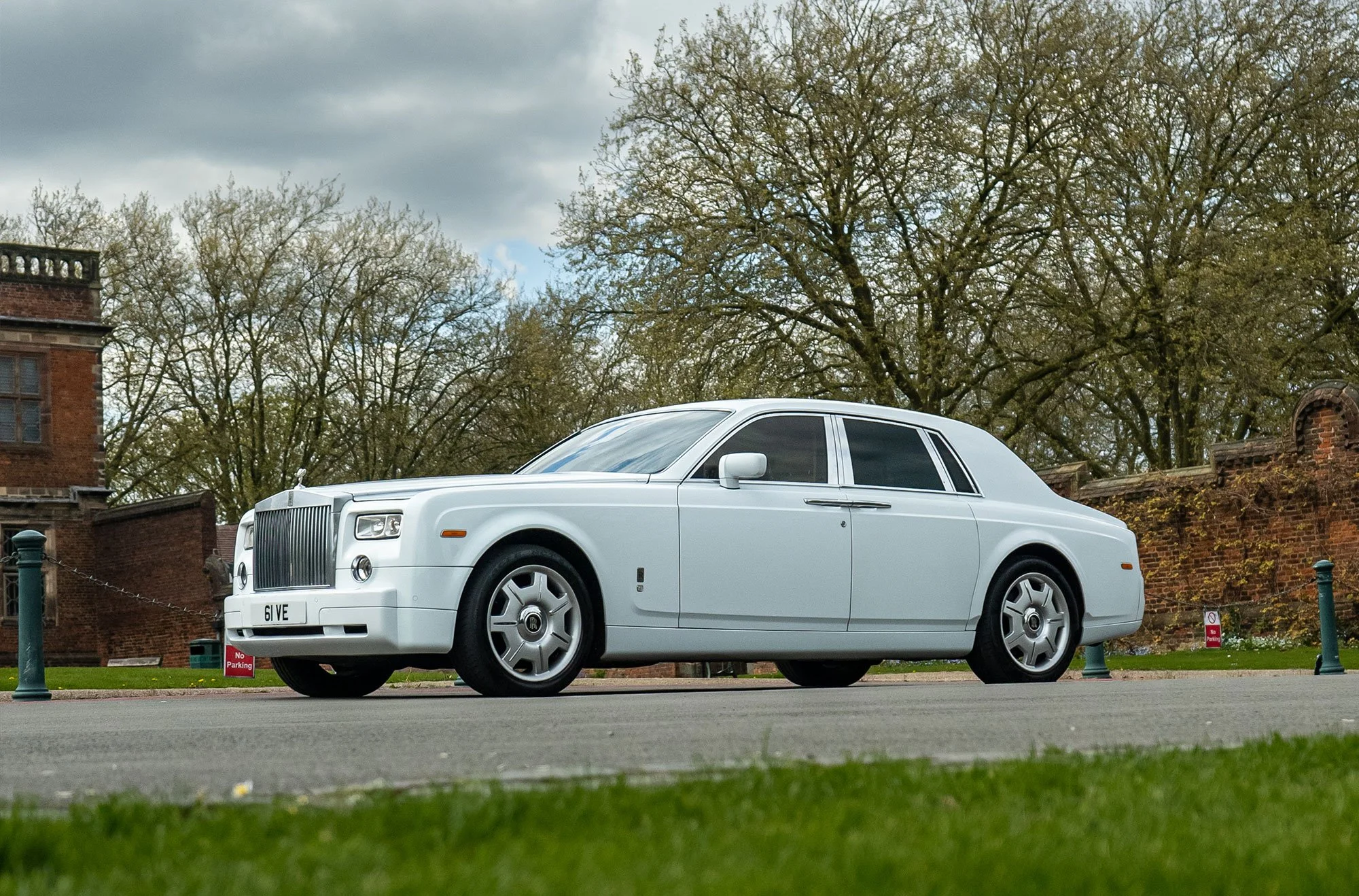 White Rolls-Royce parked outdoors with trees and brick wall in the background.