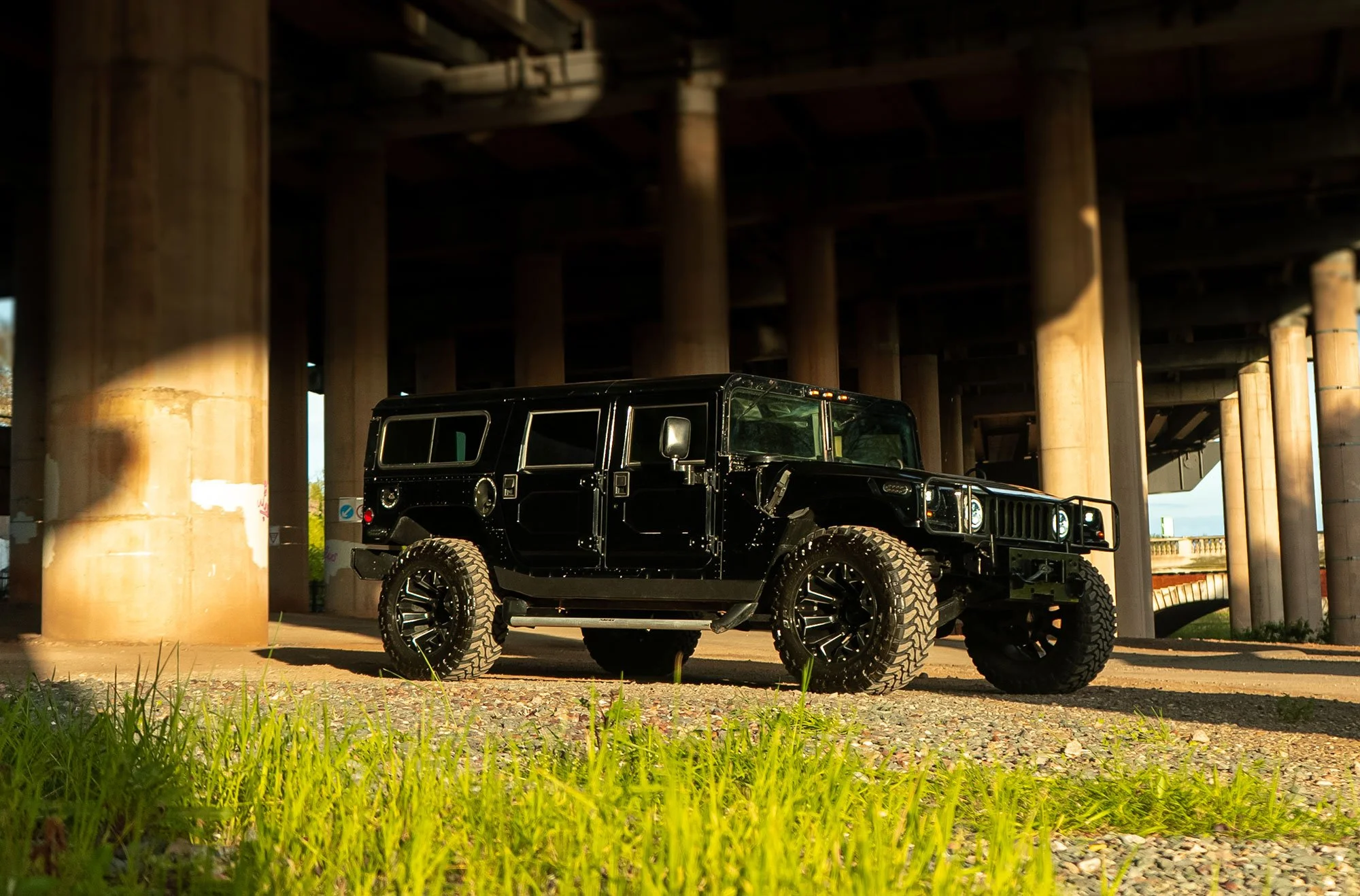 Black Hummer H1 SUV parked under a bridge with large tires, surrounded by concrete columns and grass.