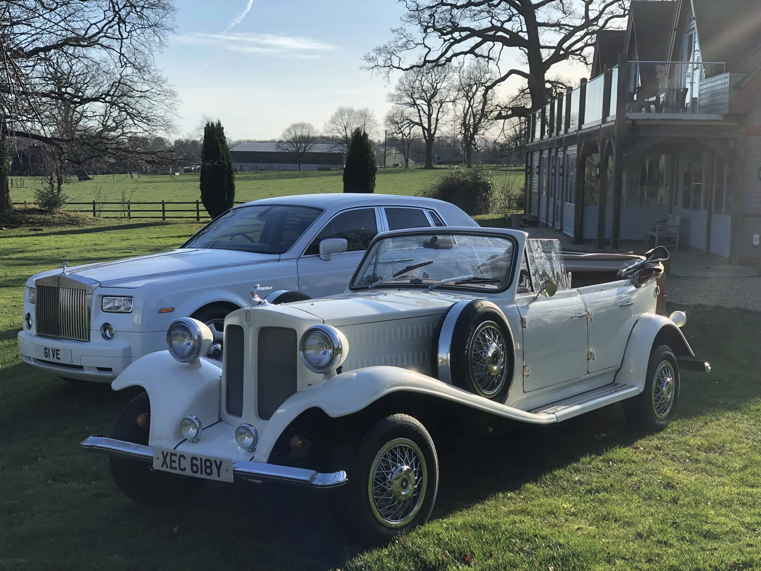 Two luxury cars parked on a grassy lawn in front of a building. The cars are a vintage convertible and a modern Rolls-Royce.