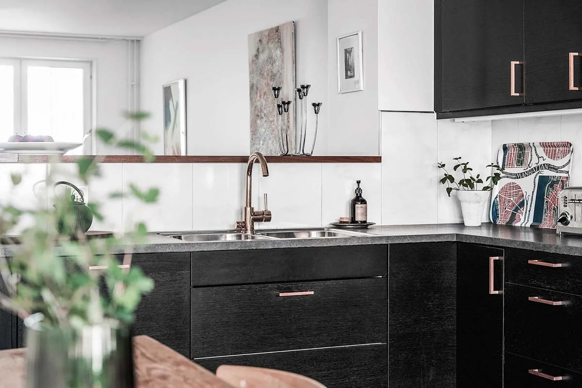 Homestyling Lidingö. Mörkt, modernt kök med mässingsbeslag och blandare, utsikt in mot vardagsrum.