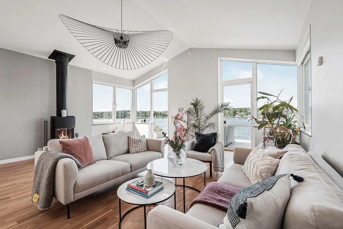 Homestyling Lidingö. Ljust vardagsrum med soffor, kamin, stora fönster och taklampa.