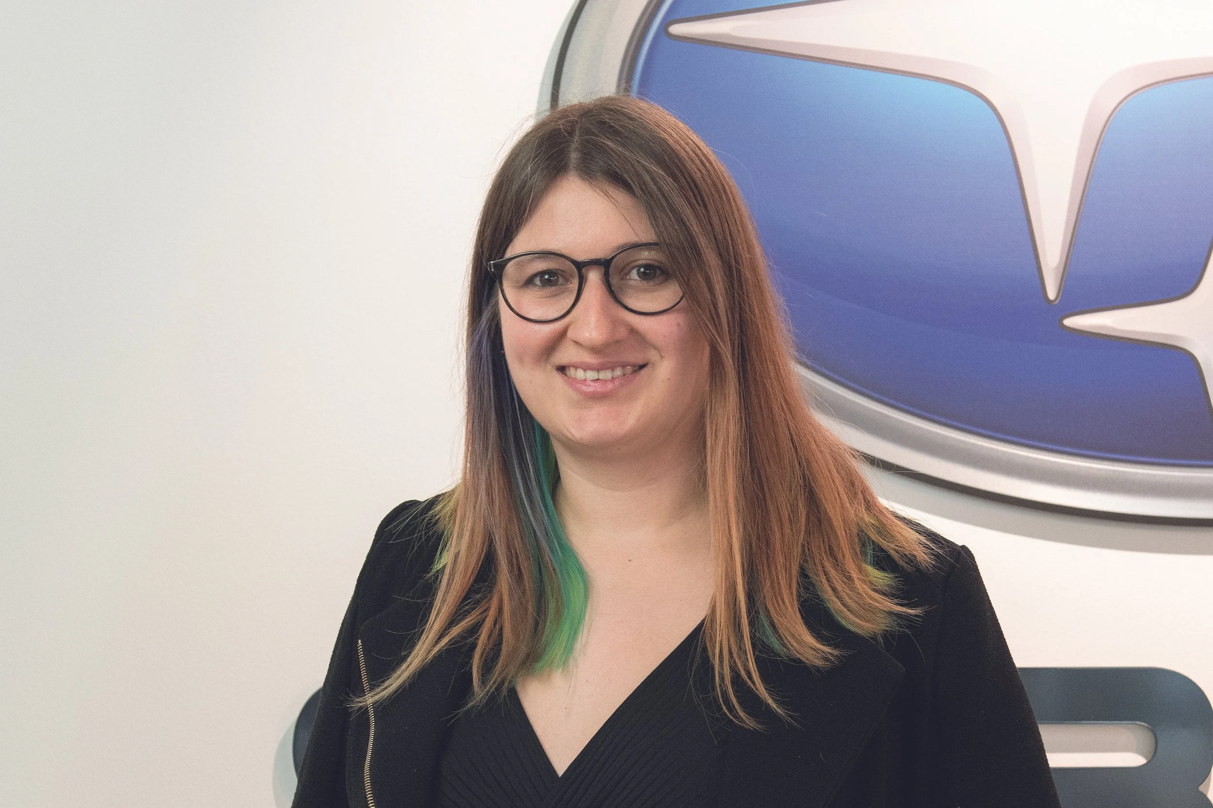 Junge Frau mit Brille und buntem Haar vor einem Tesla-Logo.