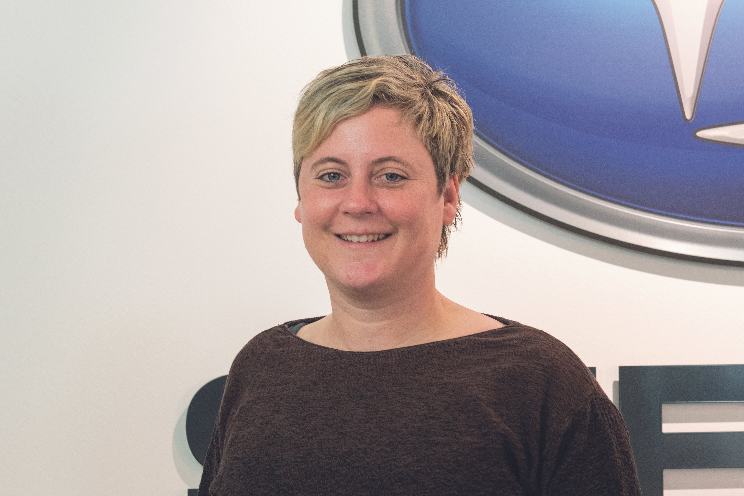 Eine lächelnde Frau mit kurzen, hellblonden Haaren vor einer weißen Wand mit einem großen blauen und silbernen Logo.