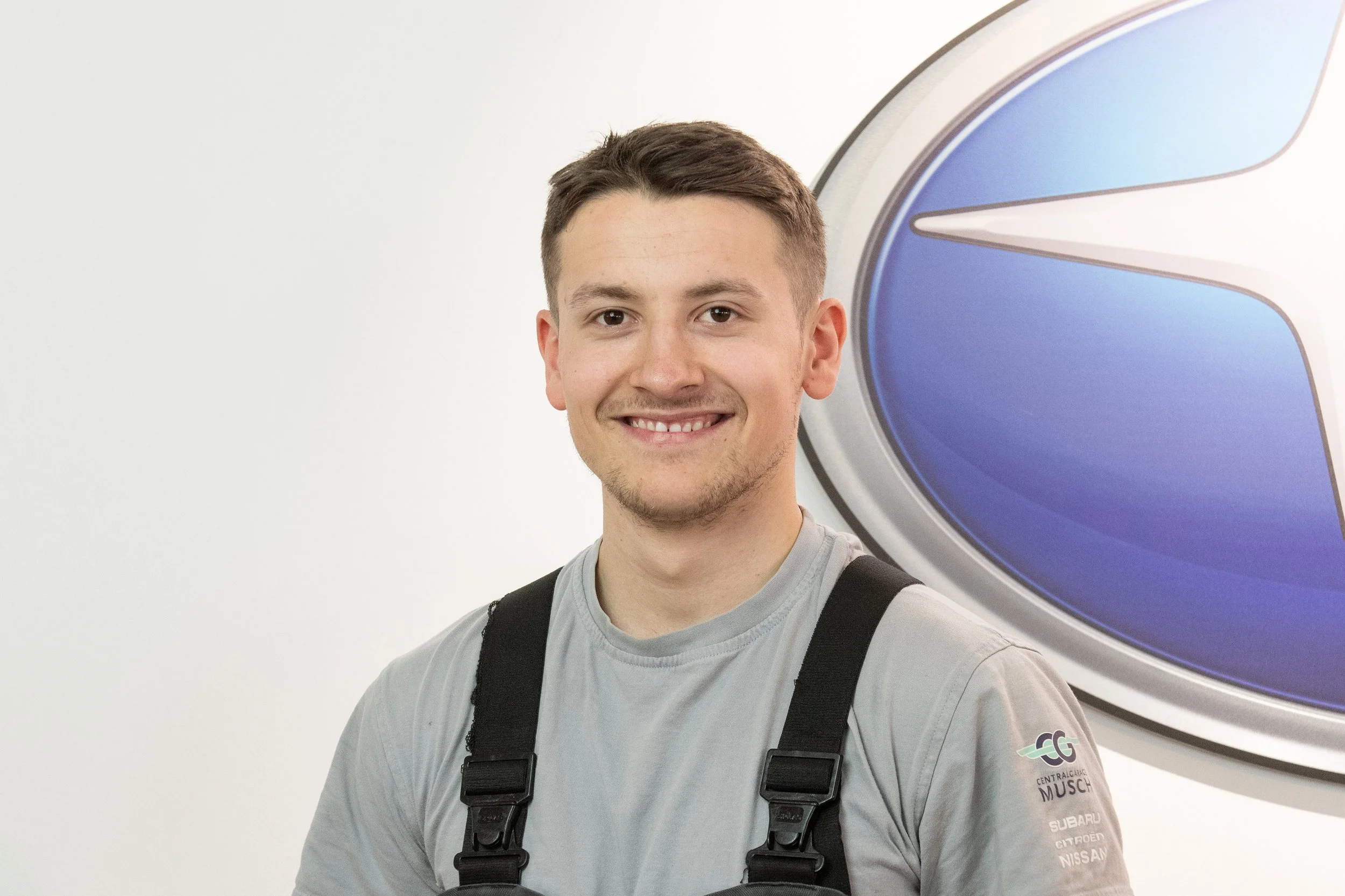 Lächelnder junger Mann trägt graues T-Shirt und schwarze Träger, steht vor einem blauen und silbernen Logo, vermutlich im Autohaus oder Servicecenter.