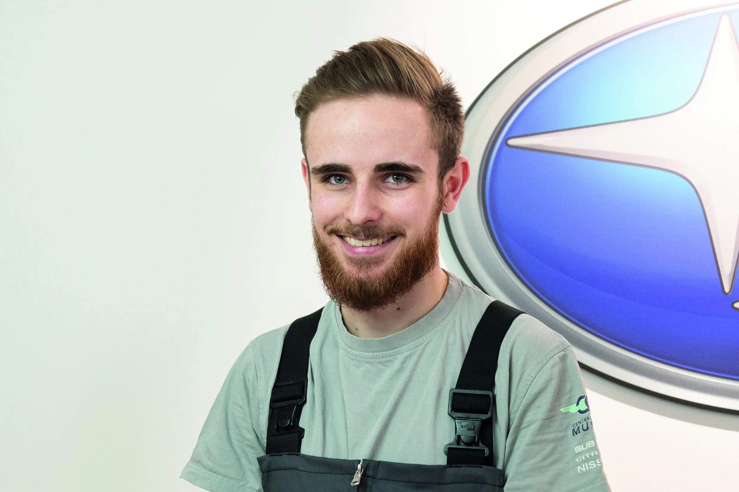 Junger Mann mit Bart und Kurzhaar frisur, trägt graues T-Shirt mit Firmenaufdruck, steht vor einem Subaru-Logo, lächelt in die Kamera.