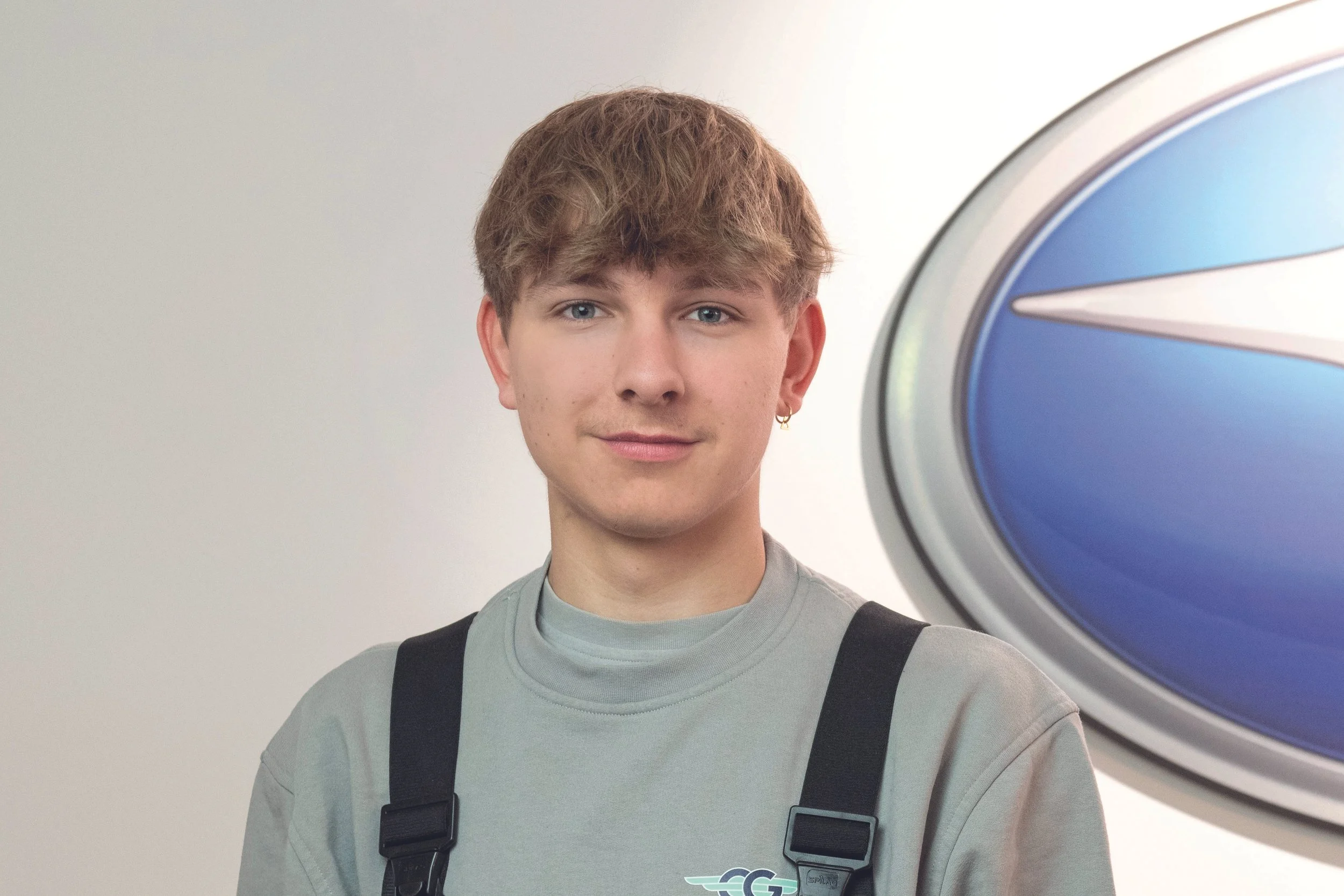 Junger Mann steht vor einer Waschanlage, trägt graues Sweatshirt und schwarze Schultergurte.