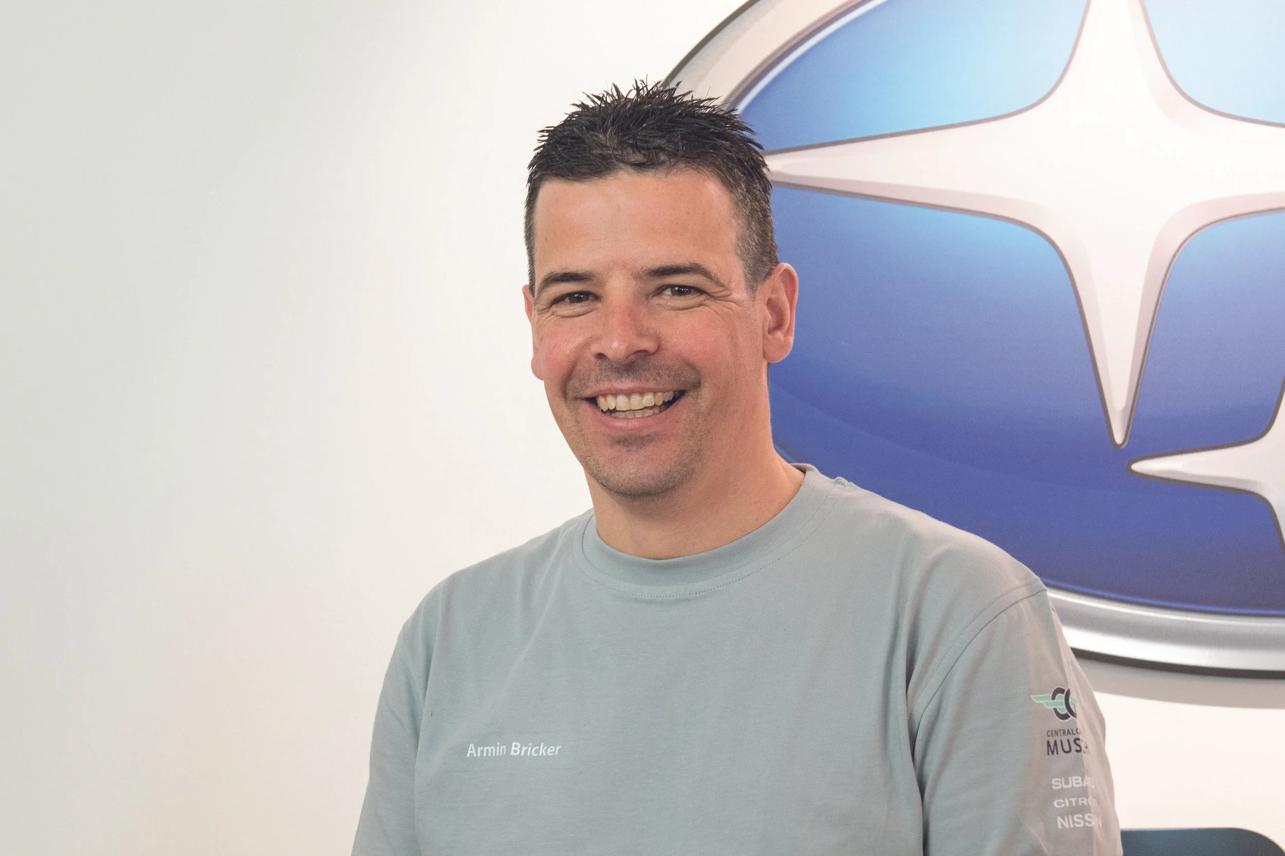Ein lächelnder Mann trägt ein hellgraues T-Shirt mit der Aufschrift 'Armin Bricker' und steht vor einer Wand mit einem großen blauen und weißen Logo.