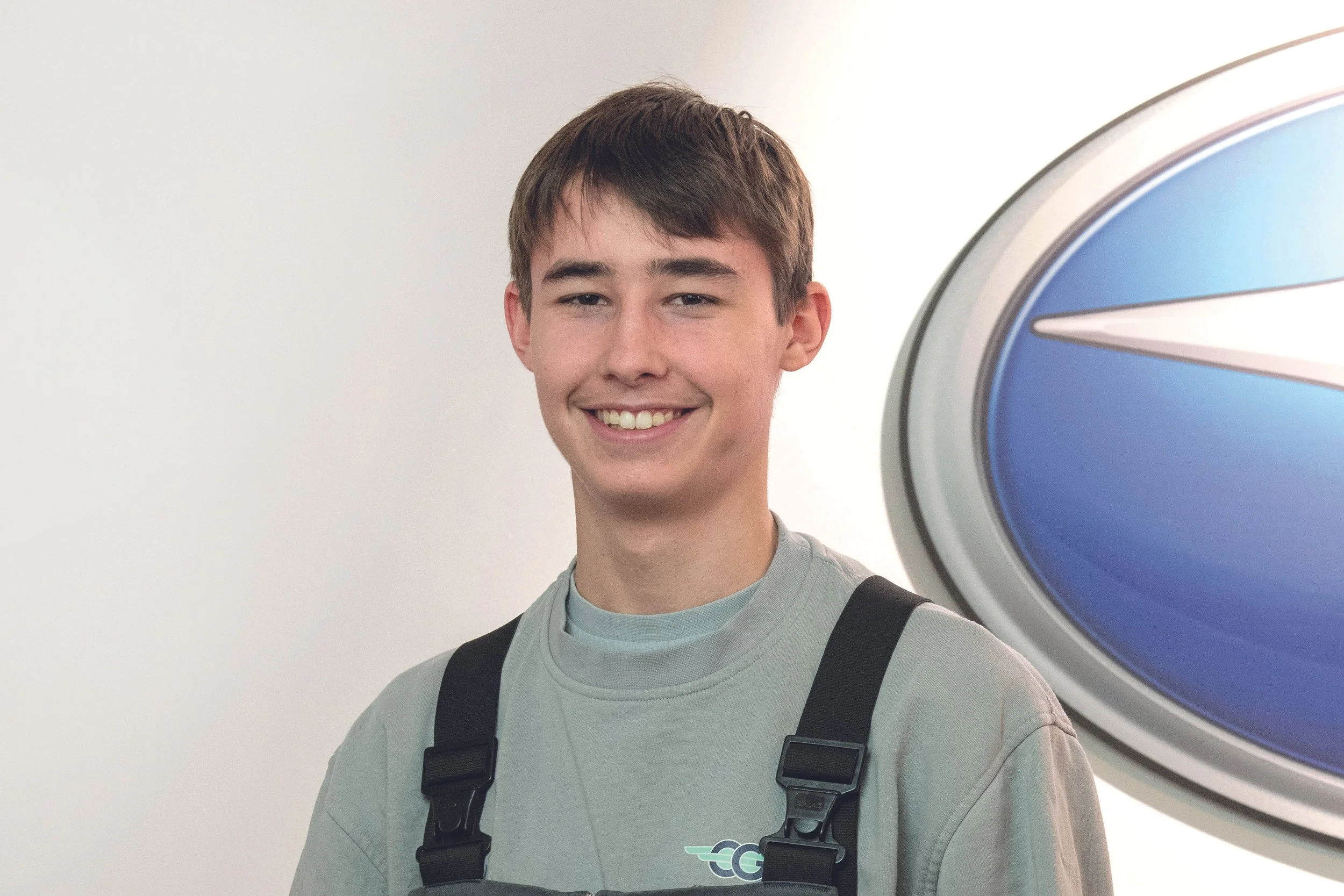 Junge männliche Person in einem grauen T-Shirt mit schwarzer Tragetasche, lächelnd vor einem blauen und silbernen Logo