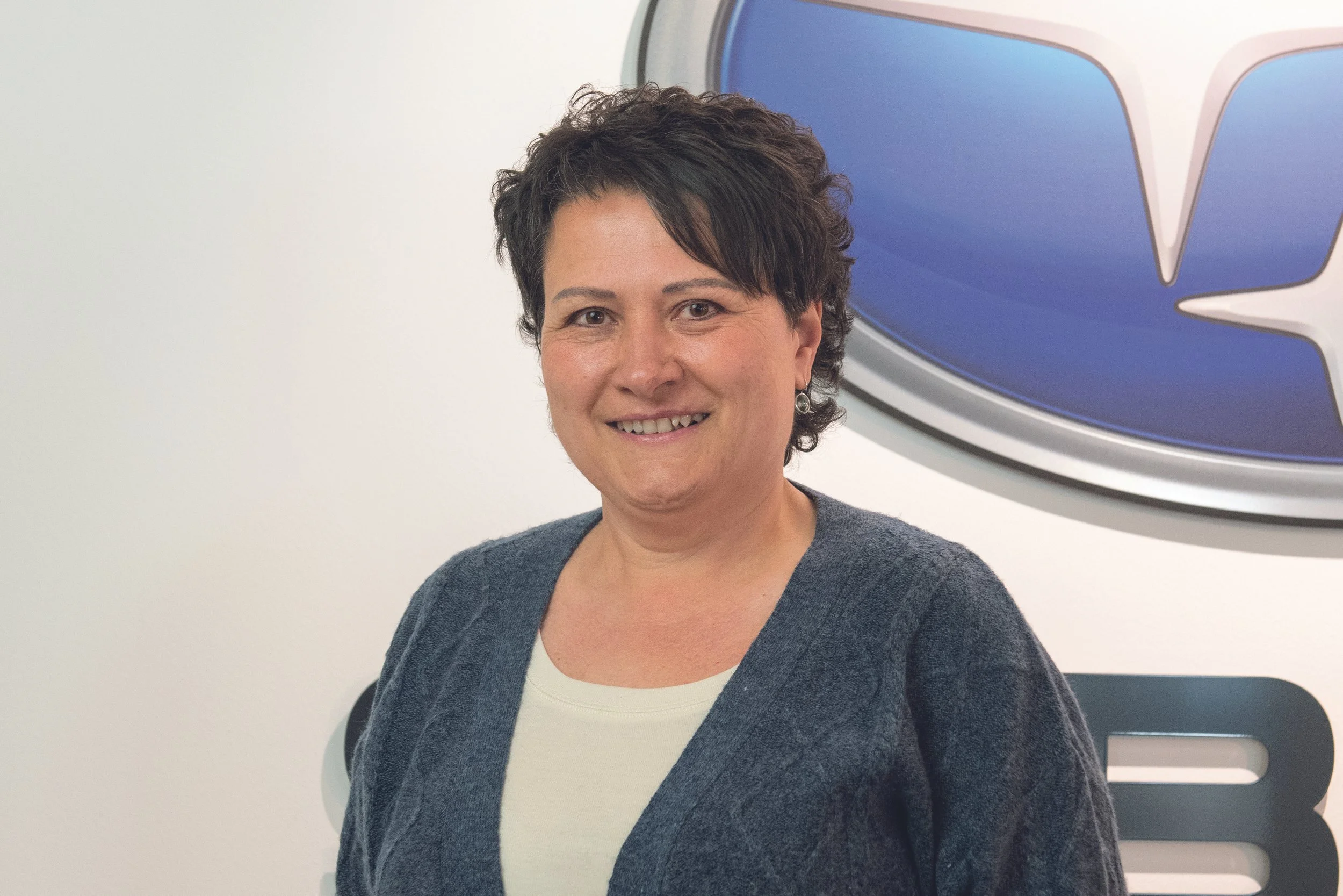 Porträt einer Frau mit kurzen dunklen Haaren, die vor einem blauen Logo steht, lächelnd.