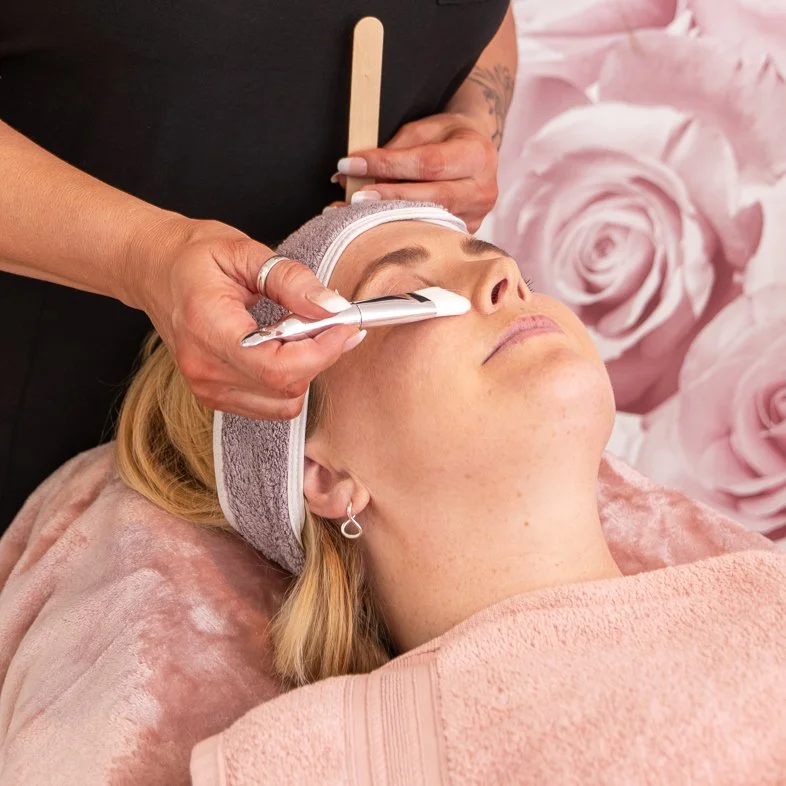 A person receiving a facial treatment in a spa setting, with a cosmetologist applying a product with a brush on their face. The individual is lying down with a headband and wrapped in a towel against a floral background.