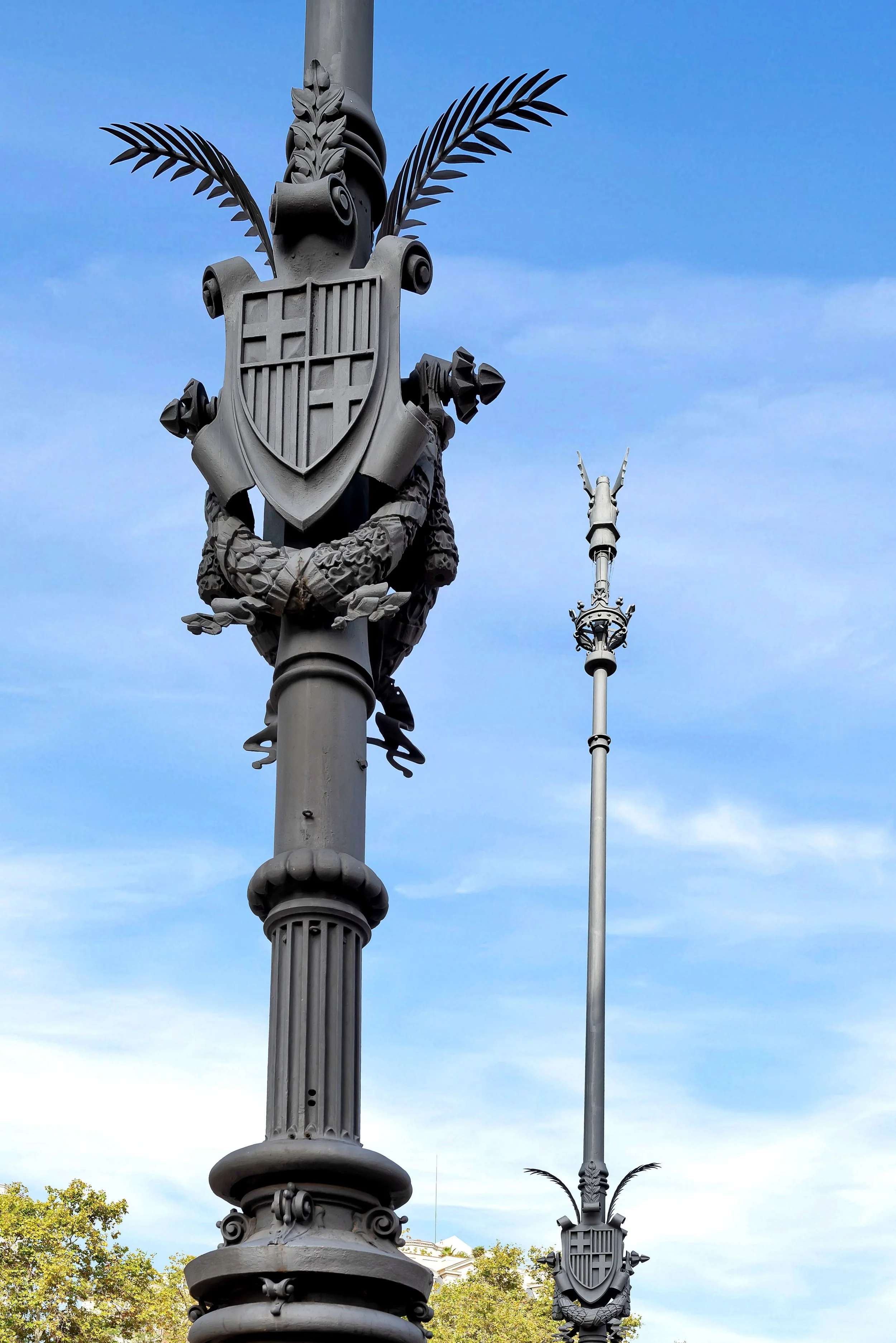 Farolas decorativas con escudos heráldicos y detalles ornamentales, en un día soleado con cielo azul y árboles en el fondo.
