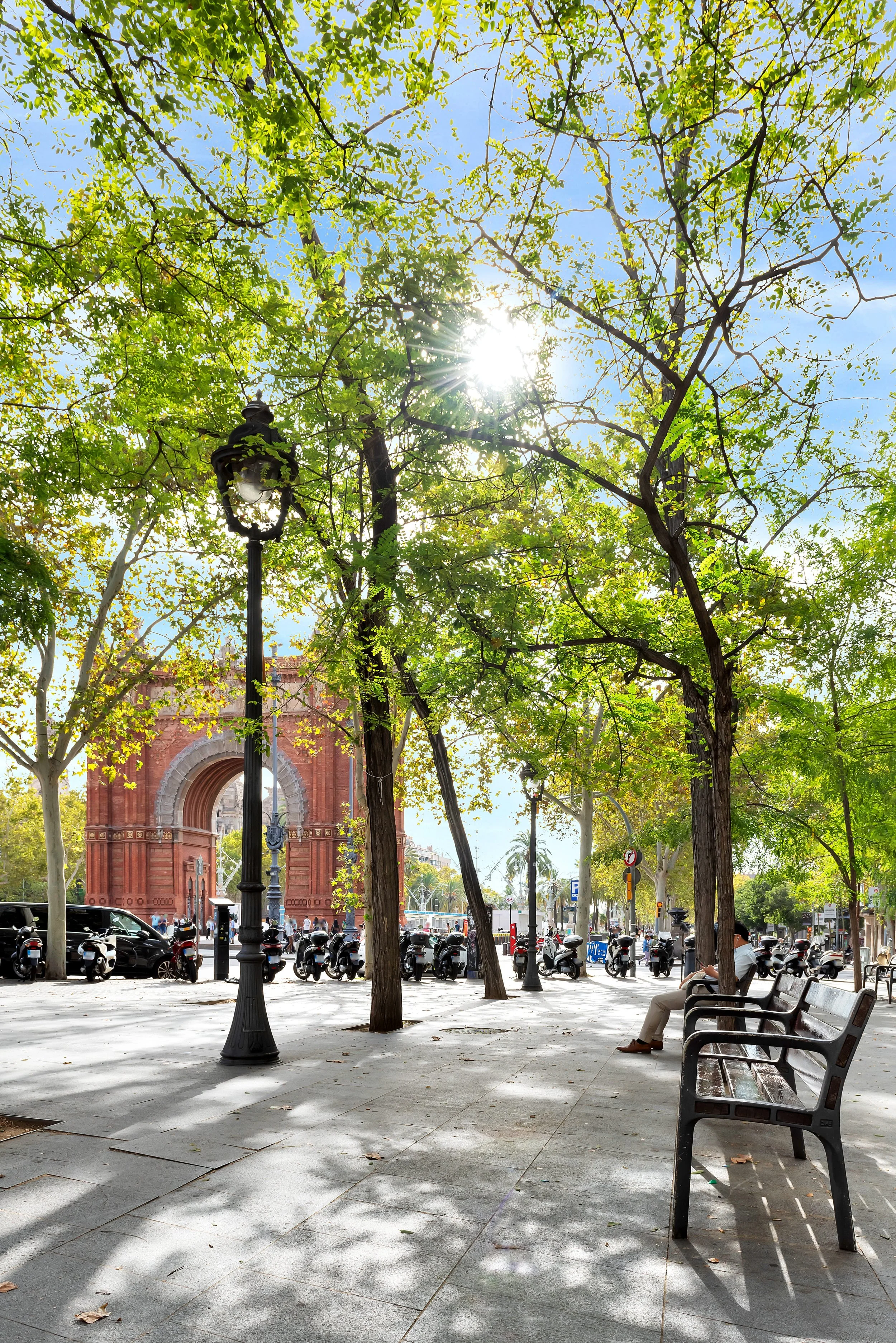Parque urbano con árboles, bancos, farola y muchas motos en la calle, con Arco de Triunfo en el fondo, en un día soleado con cielo despejado.