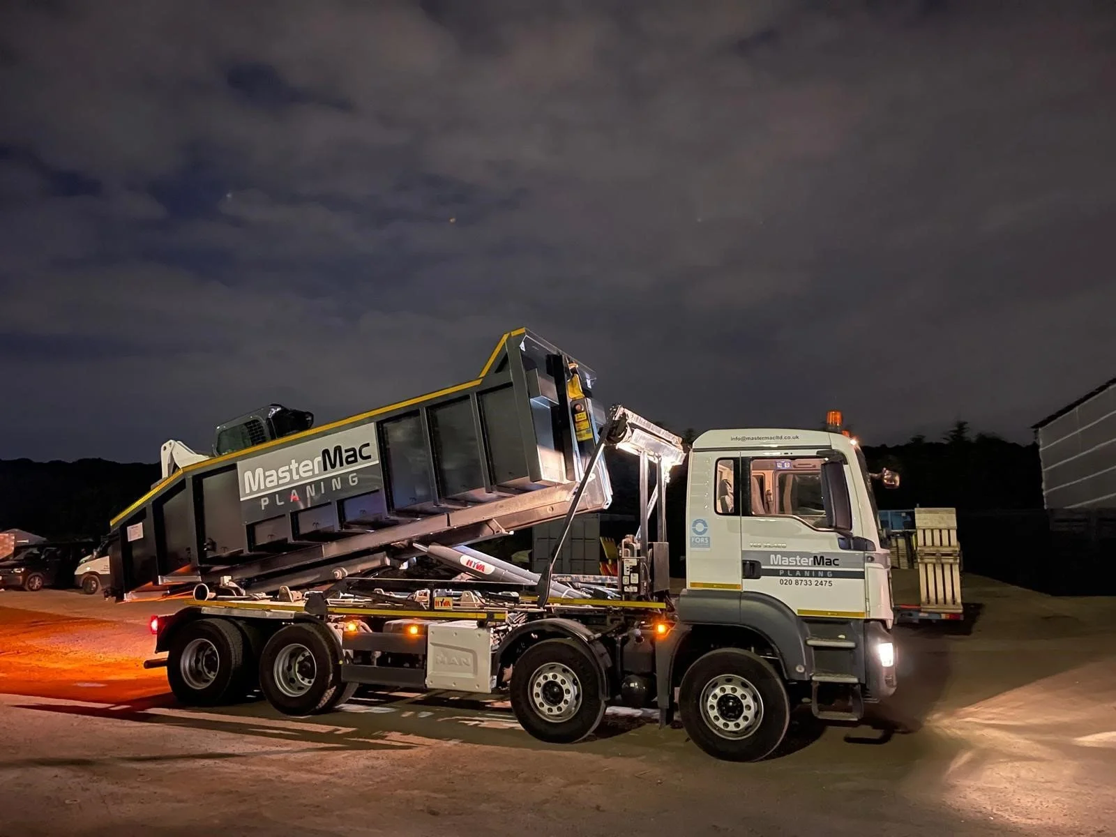 A flatbed truck with a hydraulic lift is transporting a large black and yellow container labeled 'MasterMac Planning' at night, with a dark sky and some clouds overhead.