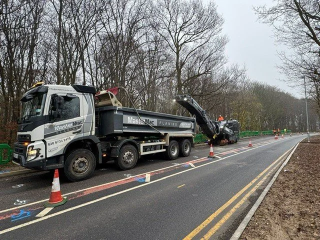 Two large road maintenance trucks are stopped on the side of a two-lane road with orange cones placed around them. The truck in the front has its hood open, and workers are visible in the background. The scene is set on a cloudy day with leafless trees lining the roadside.