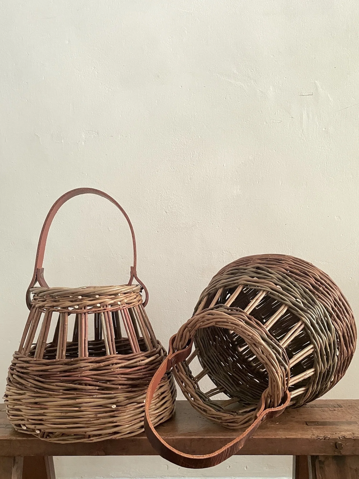 Two woven wicker baskets, one upright with a handle and one on its side, placed on a wooden surface against a plain beige wall.
