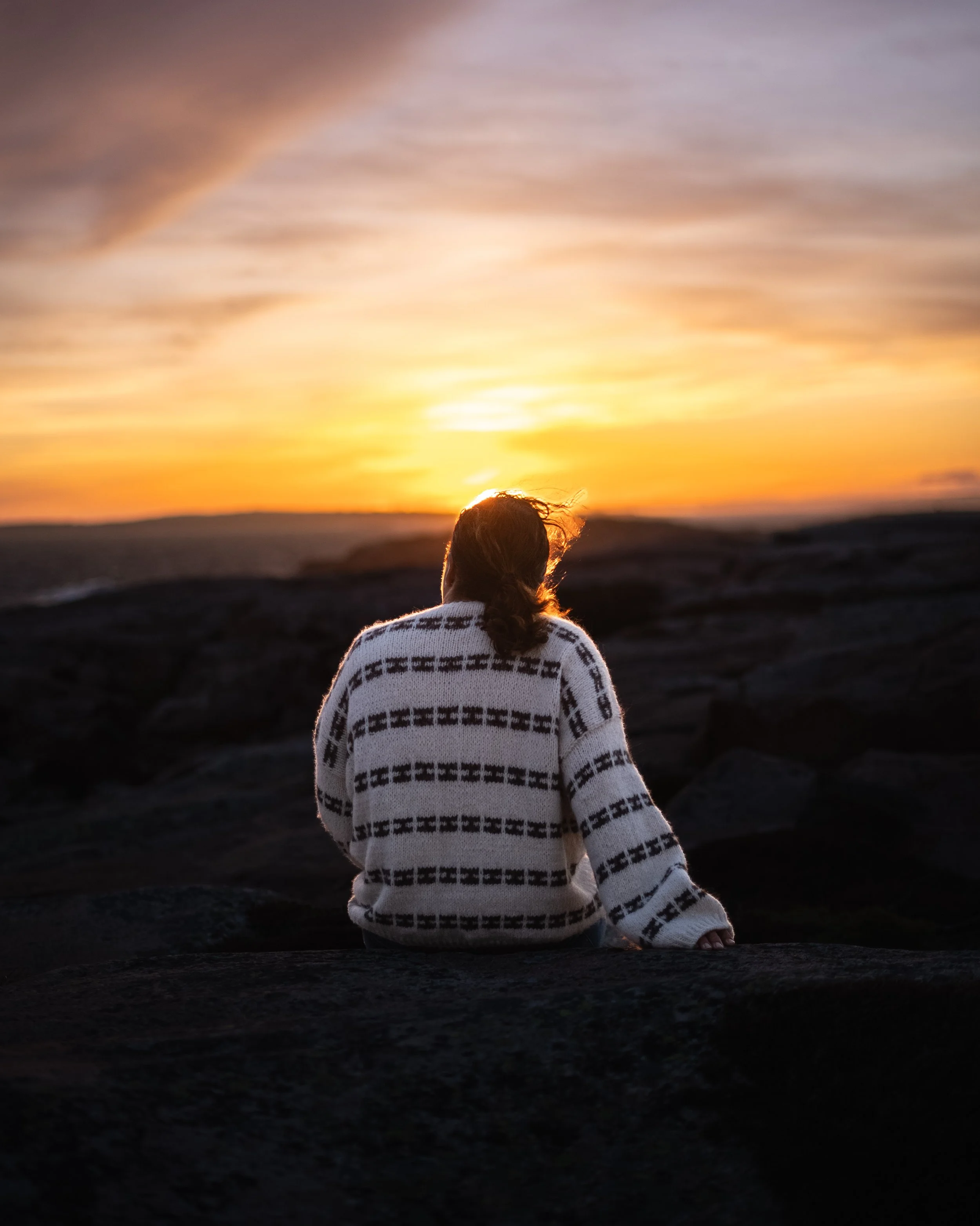 En person med langt hår i en flettet hestehale, iført en hvit genser med sorte mønstre, sitter på steiner og ser på en solnedgang over en åpen landskap.