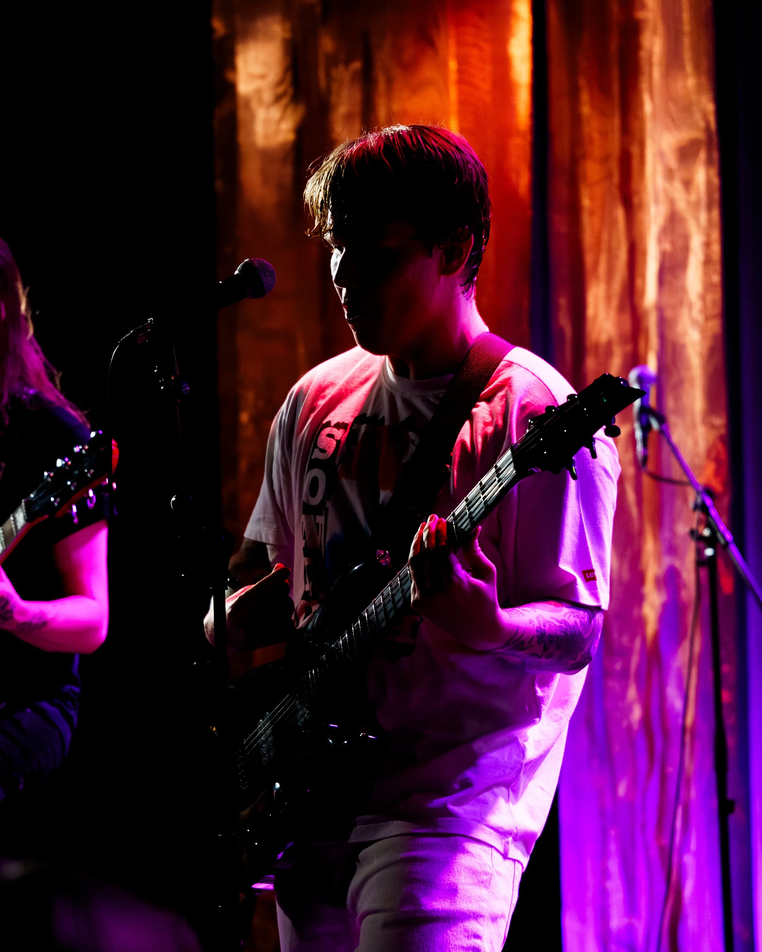 En person spiller gitar på en scene med fargede lys og brune gardiner i bakgrunnen.