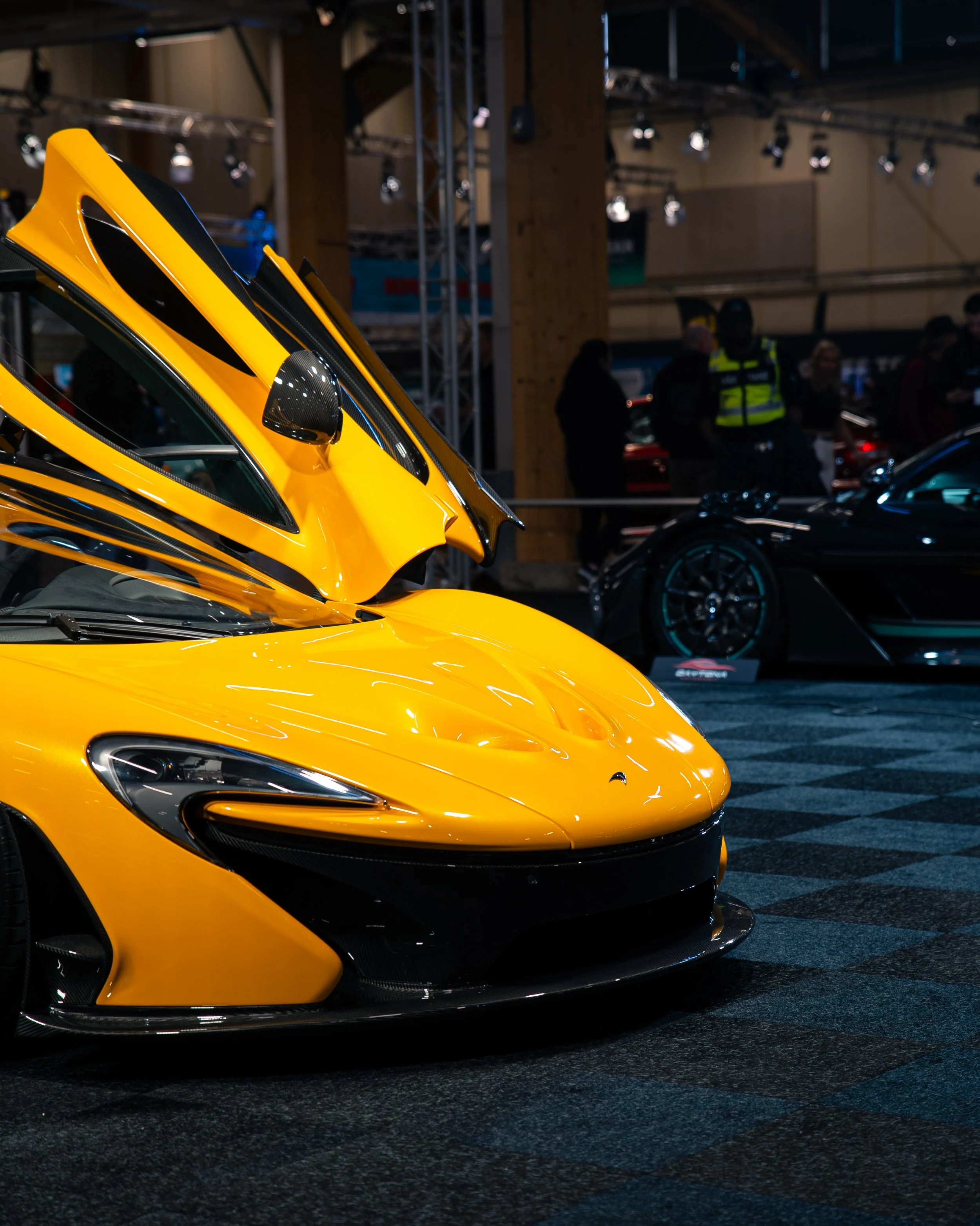 Gul sportsbil med åpne panser på en utstilling, med andre biler og mennesker i bakgrunnen.
